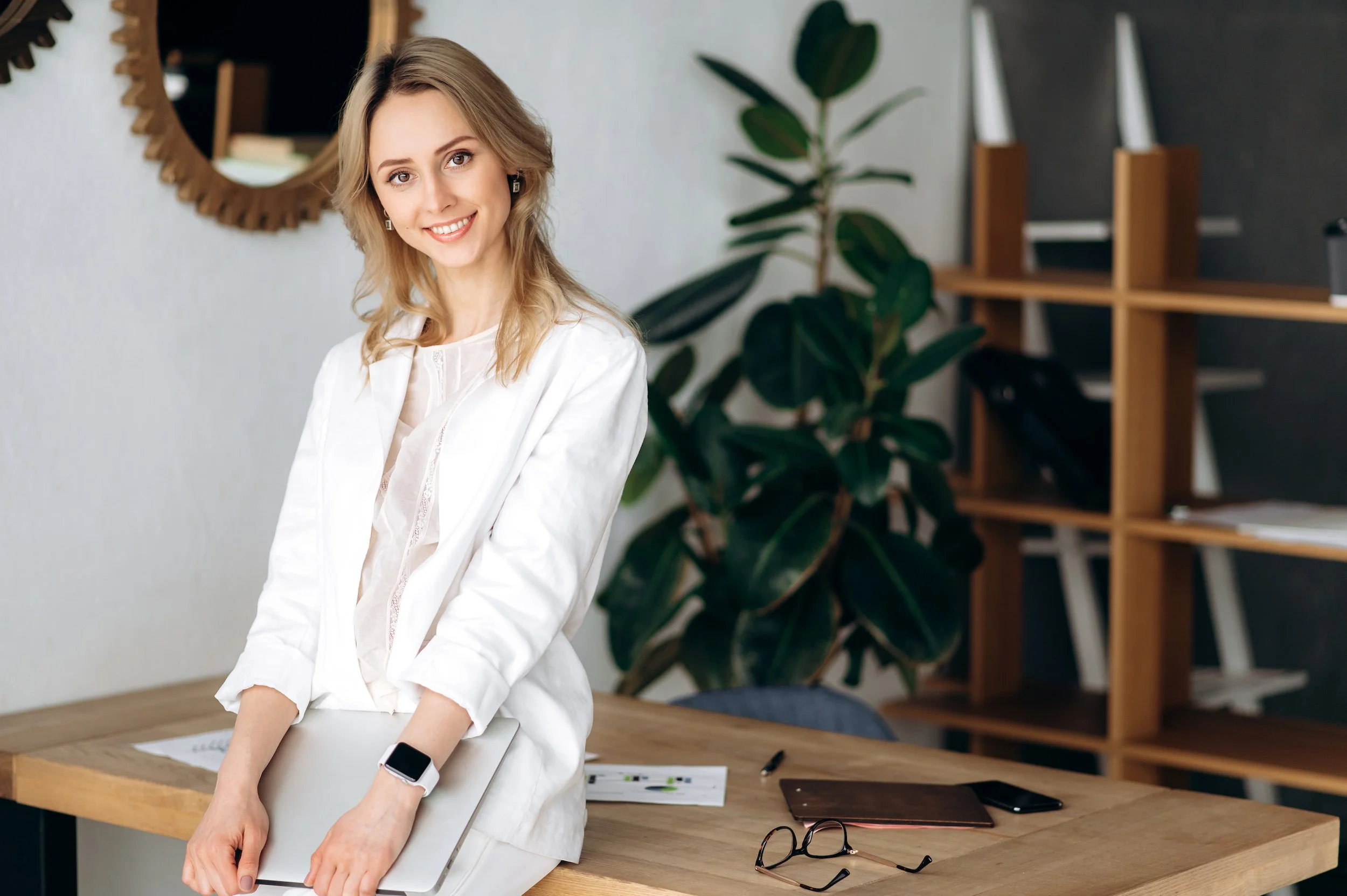 A smiling woman with blonde hair in a white blazer holding a silver laptop on a wooden table in a modern room with a mirror, plant, and shelf.