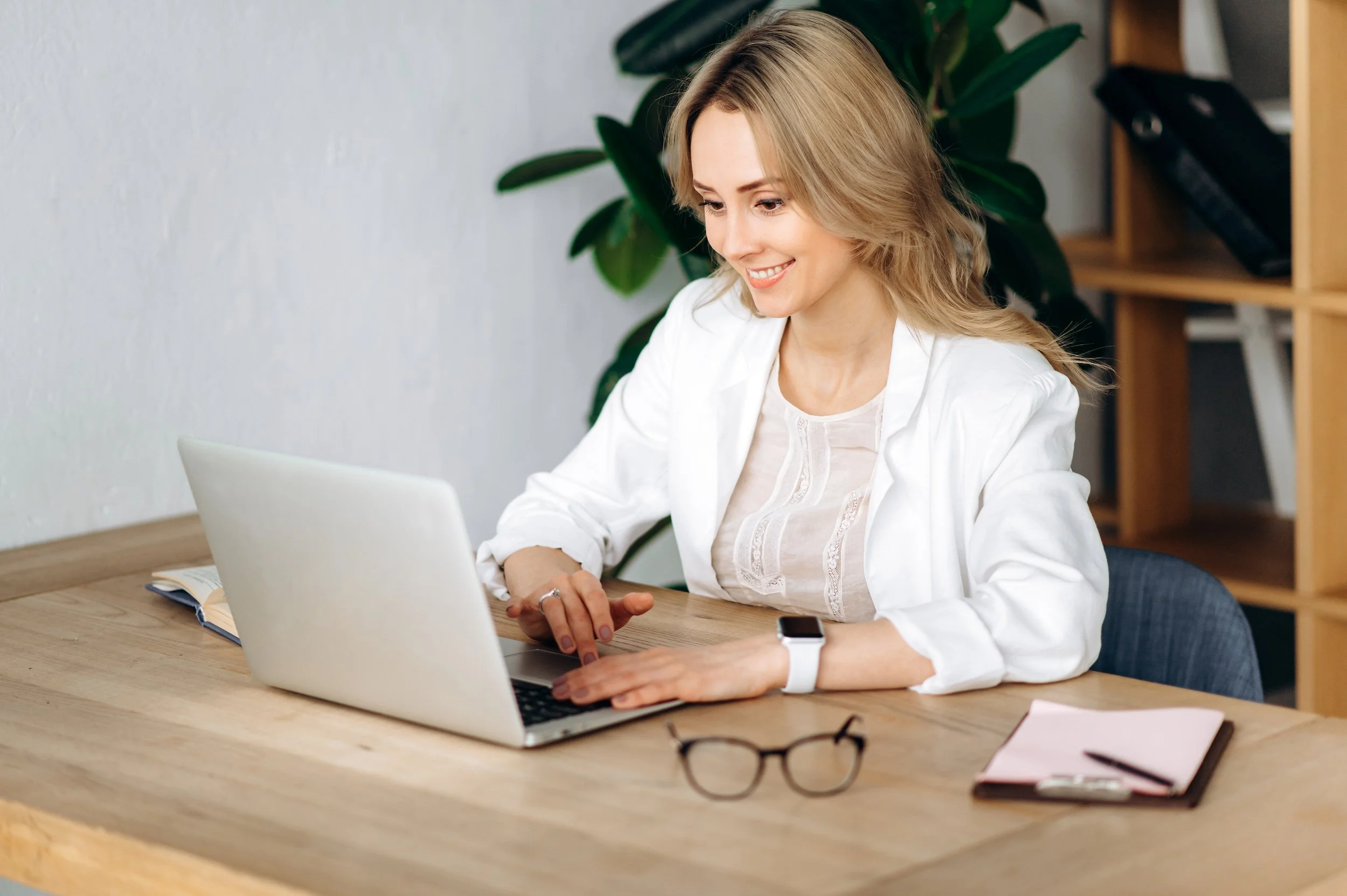 A woman with blonde hair smiling while working on a laptop at a wooden desk with glasses, a notebook, and a pen. She is wearing a white shirt and a smartwatch. Behind her are green plants and shelves with books and a black bag.