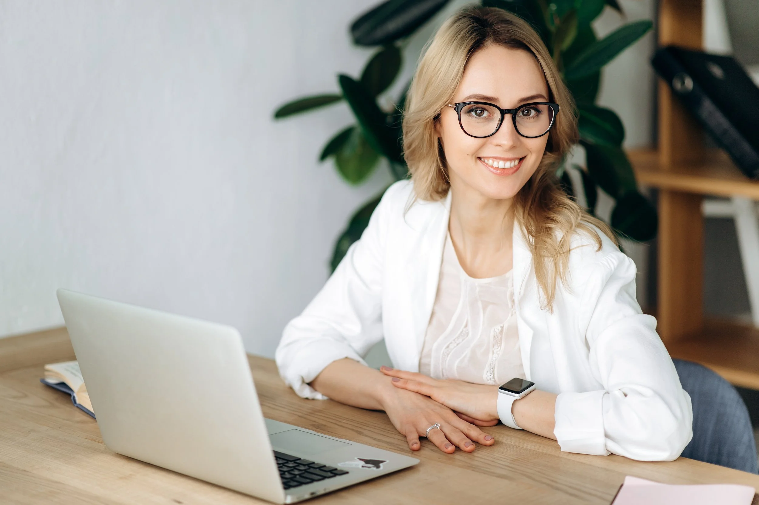 A woman with blonde hair, wearing glasses and a white blazer, smiling and sitting at a wooden desk with a laptop, notebook, and smartwatch in a bright office space with green plants in the background.