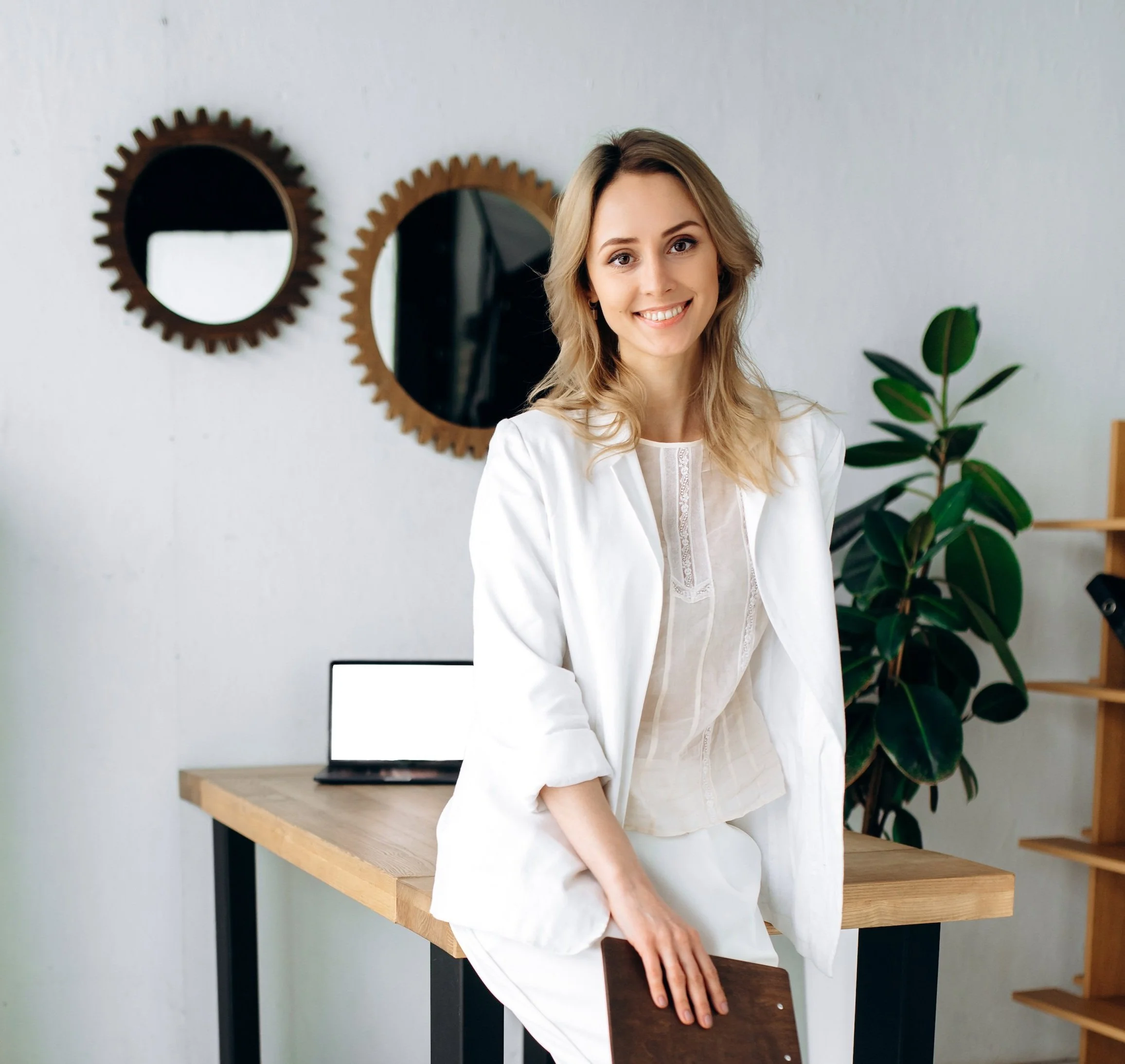 A woman with blonde hair wearing a white blouse smiling while leaning on a wooden table in a modern room with mirrors and plants.