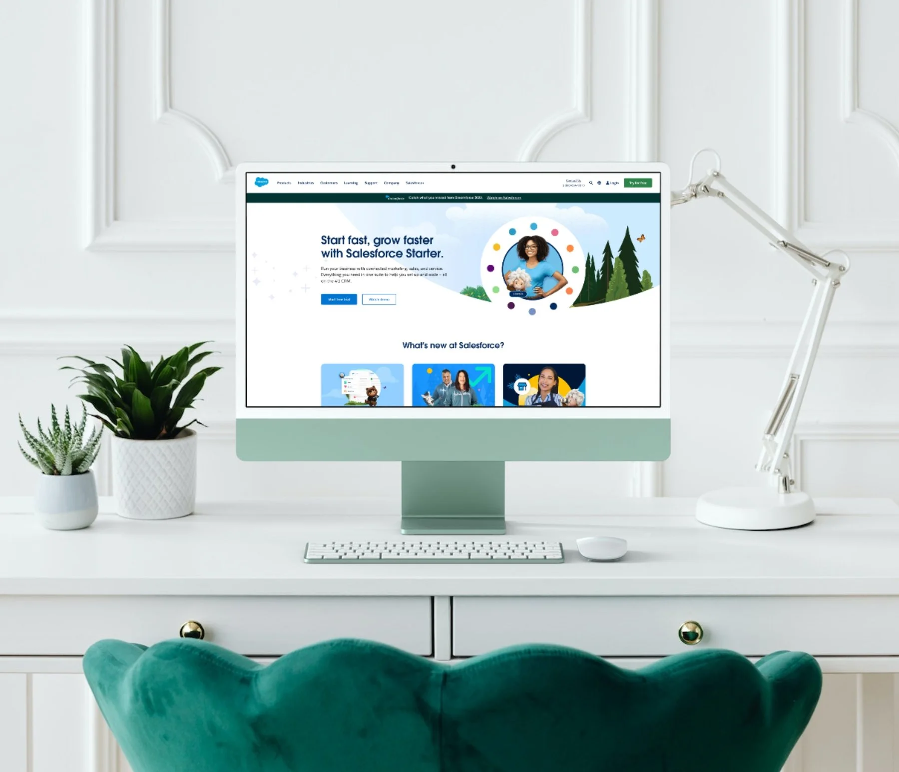 An office workspace with an Apple iMac computer displaying the Salesforce website, flanked by potted plants on the left and a white adjustable desk lamp on the right, with a plush green chair in the foreground.