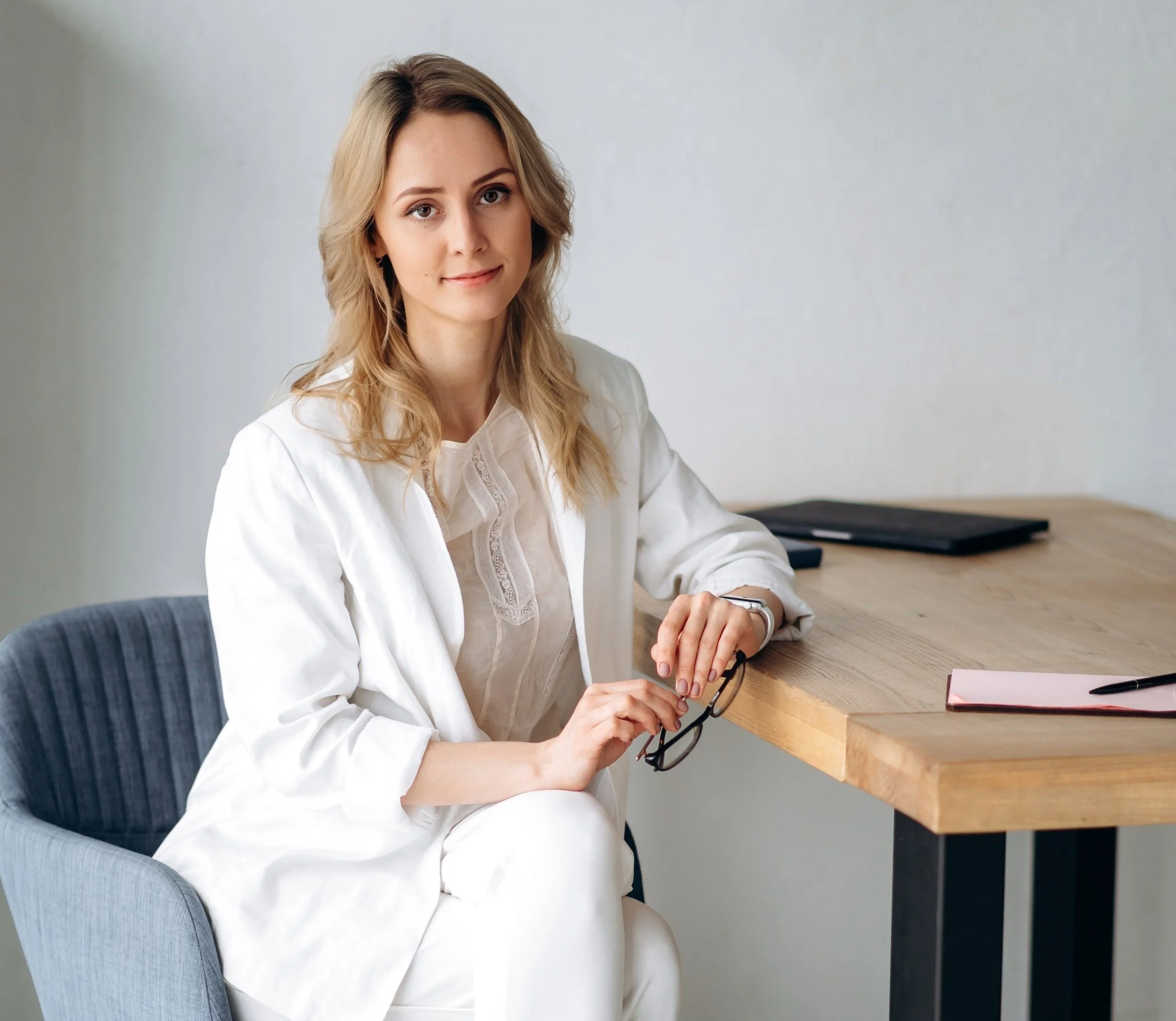 A woman with shoulder-length blond hair, wearing a white blouse, sitting at a wooden table with a black folder and a pink notepad in front of her. She is holding a pair of glasses in her hands and looking at the camera with a slight smile. The background is a plain gray wall.