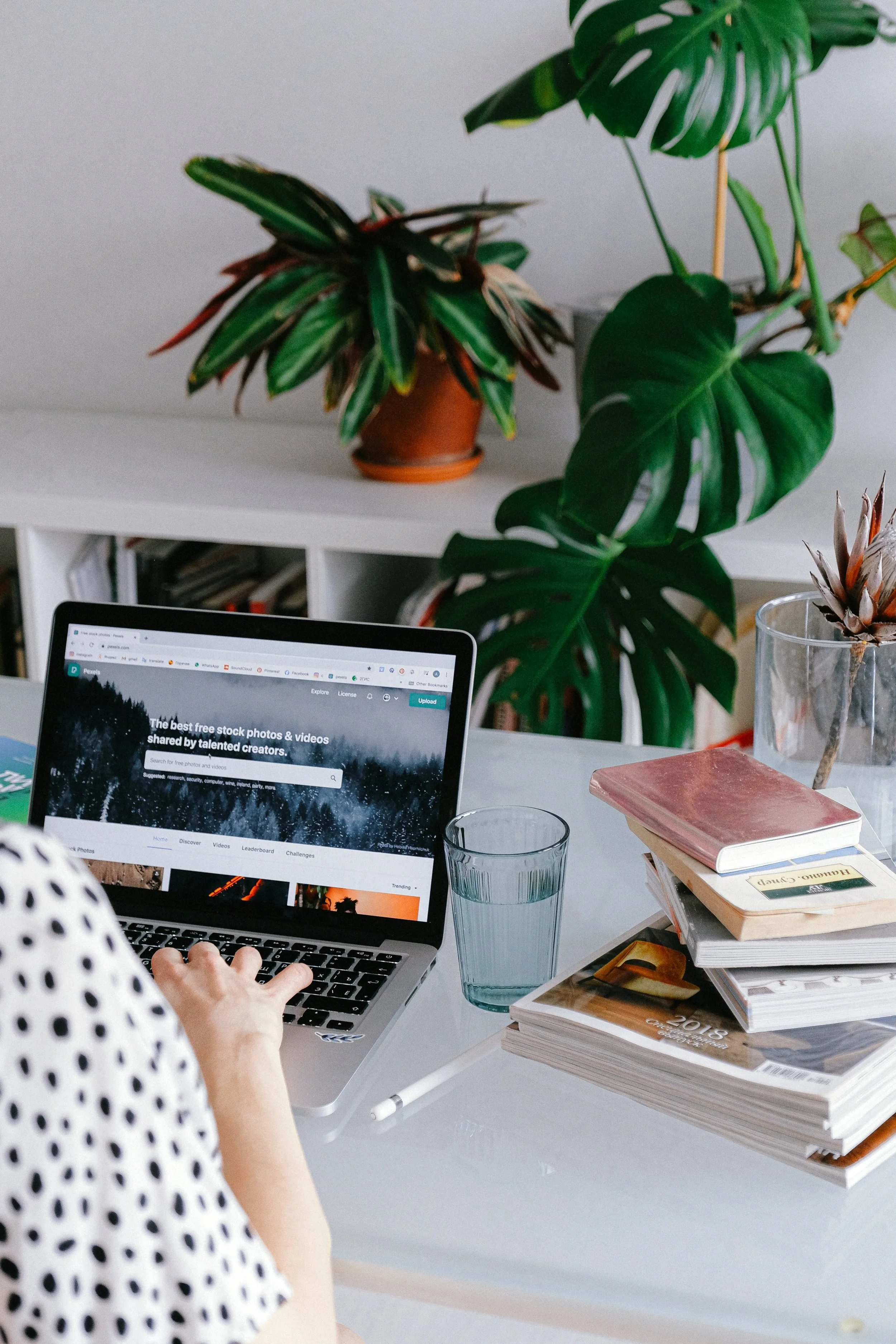 A person wearing a white shirt with black polka dots working on a laptop on a white desk. There are plants behind the desk, a glass of water, and a stack of books including a pink book and a 2018 planner.