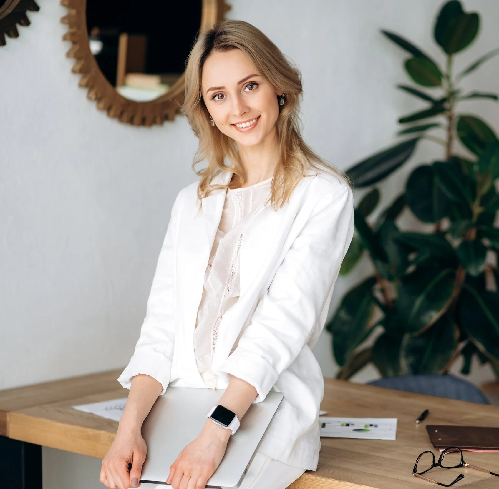 A young woman with blonde hair smiling, wearing a white blazer, holding a silver laptop, standing in an office next to a desk, with a round mirror and a large green plant in the background.