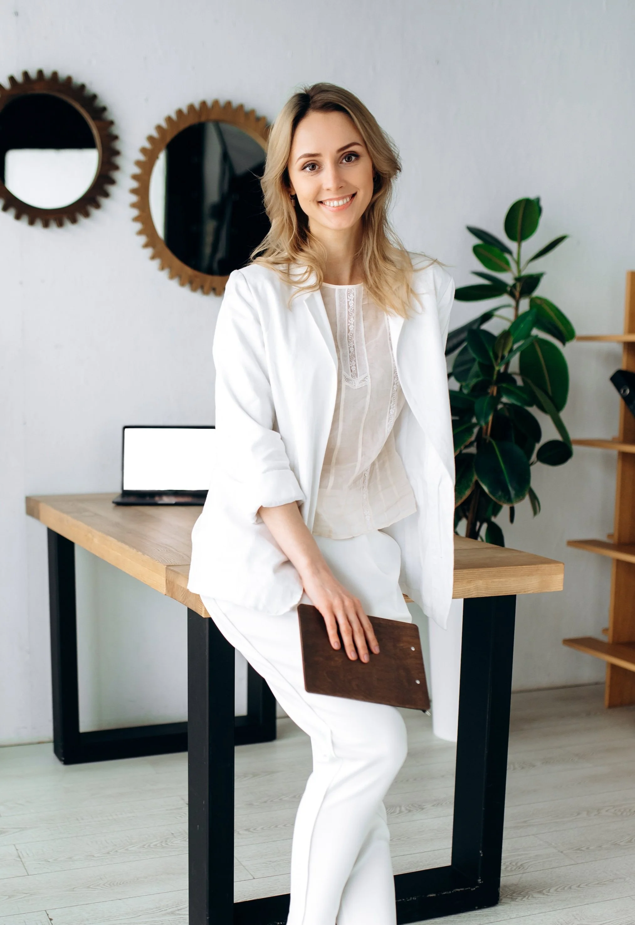 A woman with blonde hair wearing a white blazer and white pants, smiling and sitting on a chair in a modern office space with a wooden desk, black metal legs, a computer monitor, a large green plant, and decorative wall mirrors.