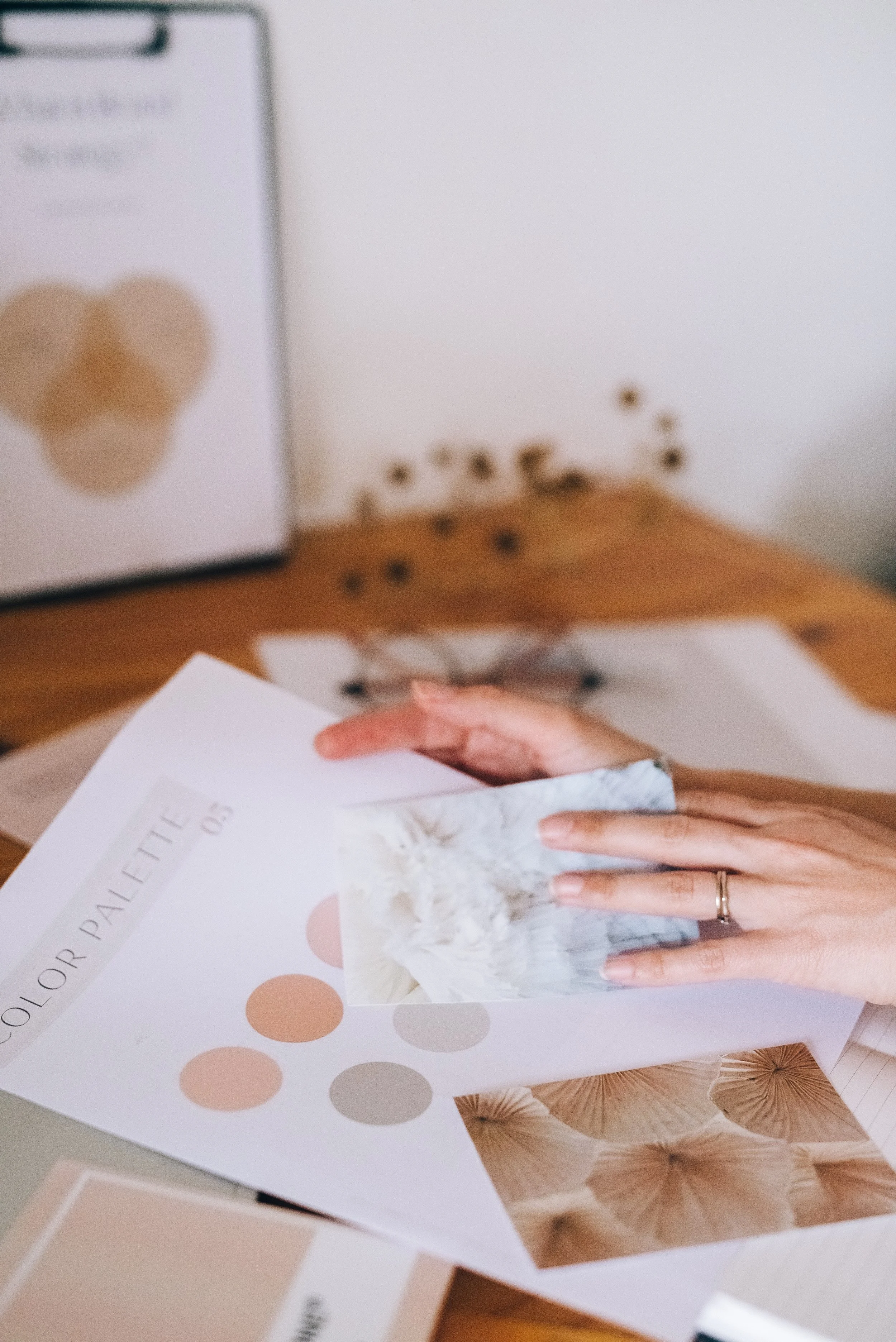 Close-up of a hand holding a brochure with color palette and design images, with a blurred background showing more design material on a table.