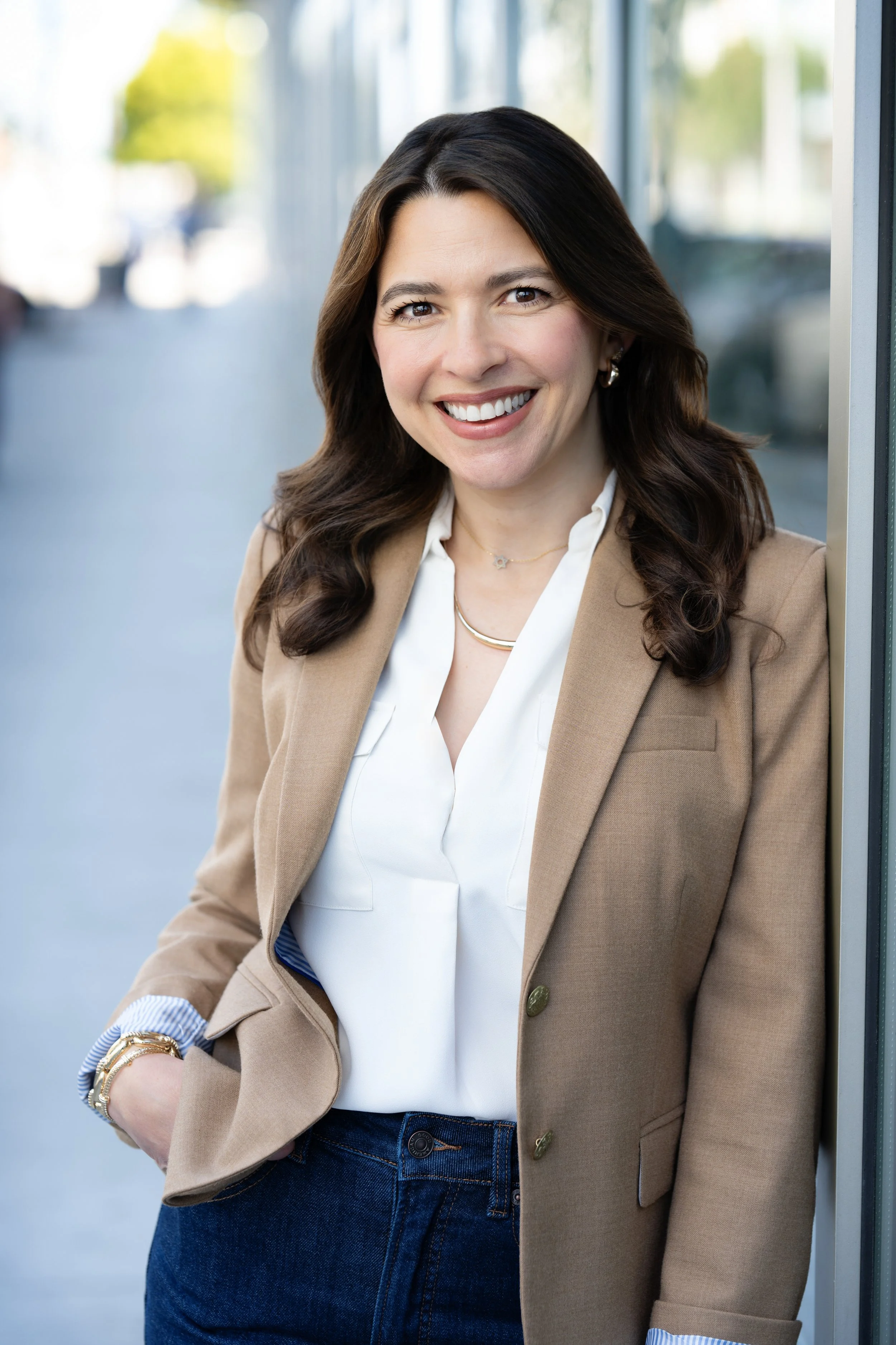 A woman with long dark hair smiling and wearing a beige blazer, white blouse, jeans, and jewelry, standing by a glass window with a blurred outdoor background.