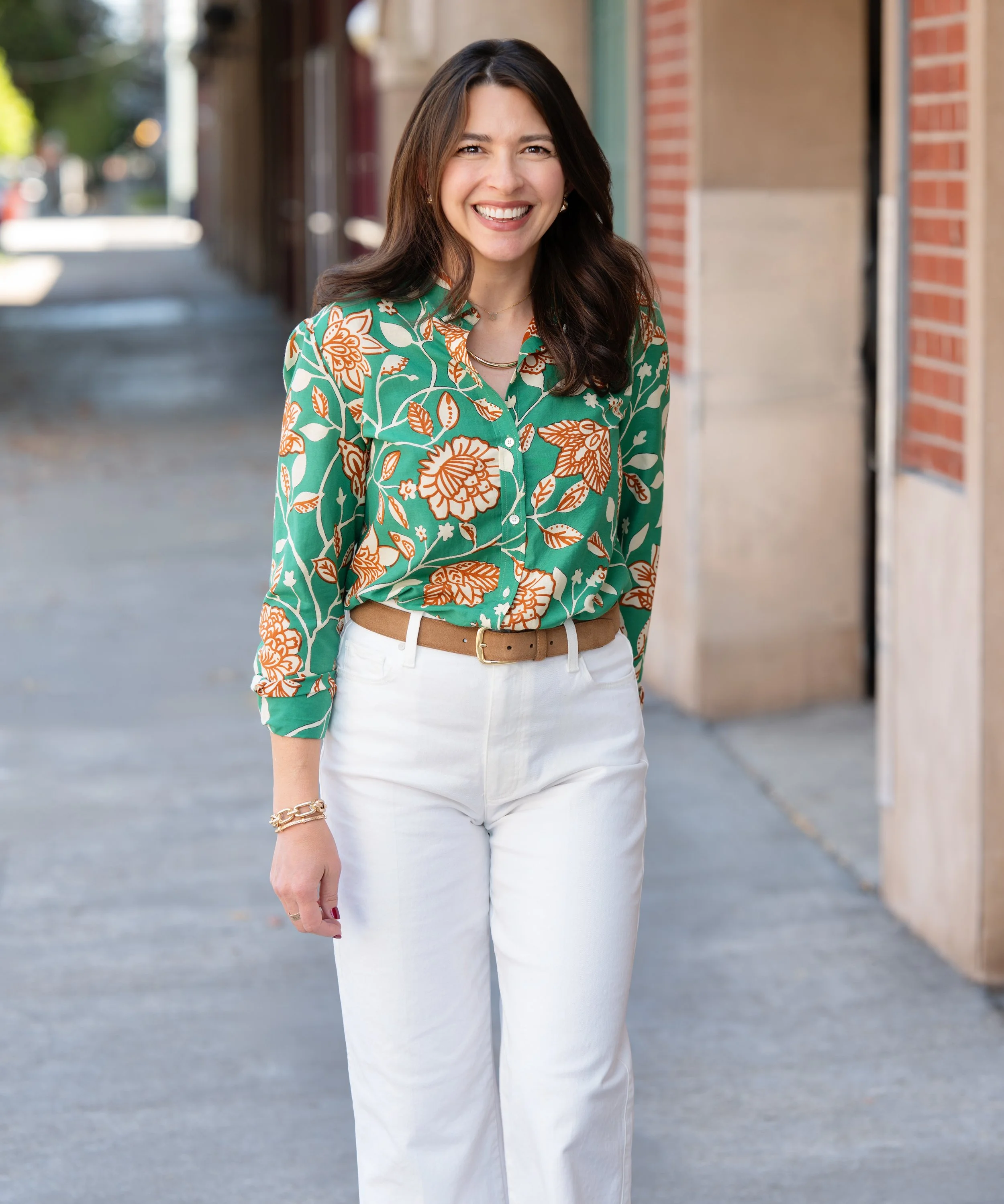 A woman with long dark hair and light skin smiling while standing outdoors on a sidewalk.