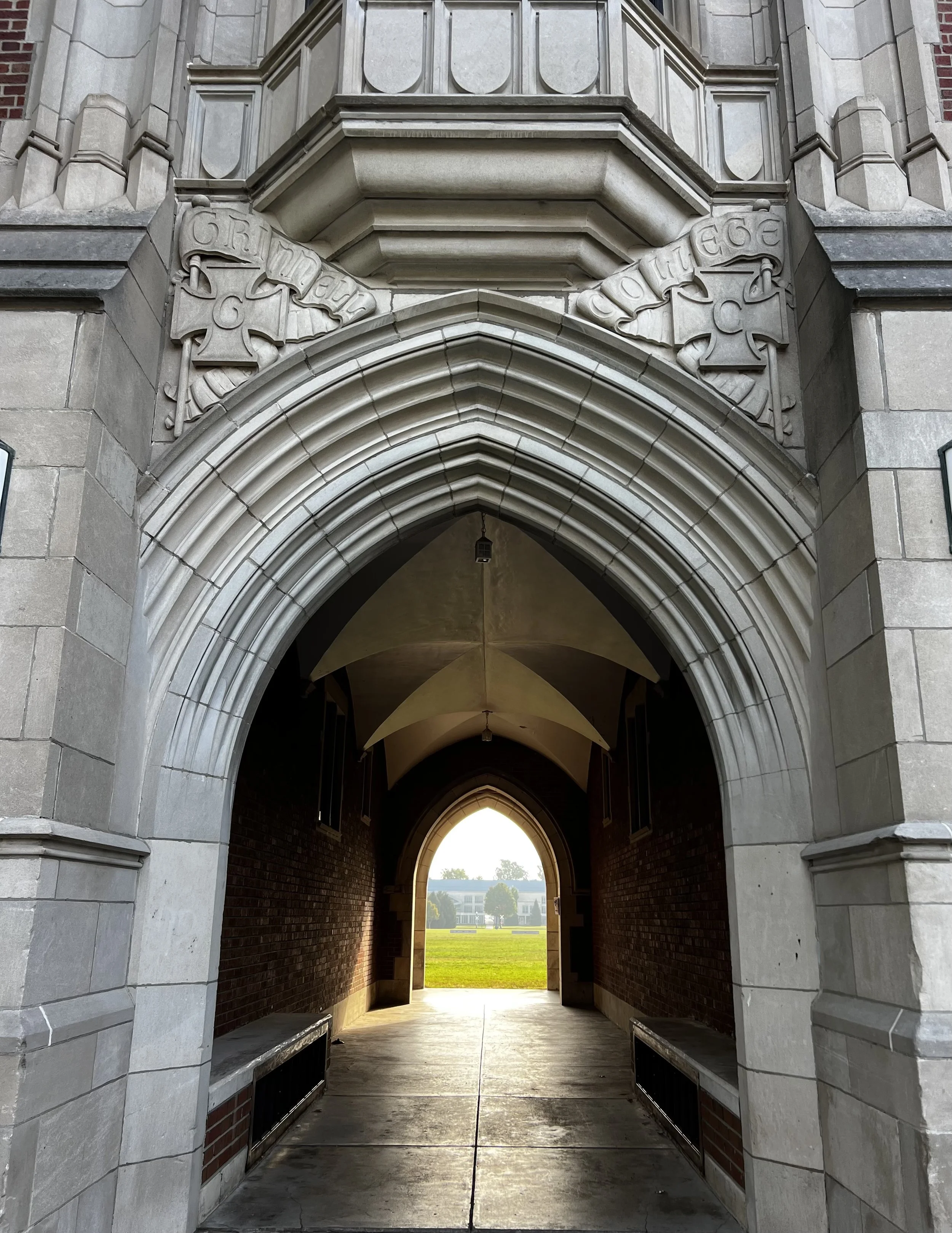 View through an arched stone gateway leading to a grassy courtyard with buildings in the distance at sunset.