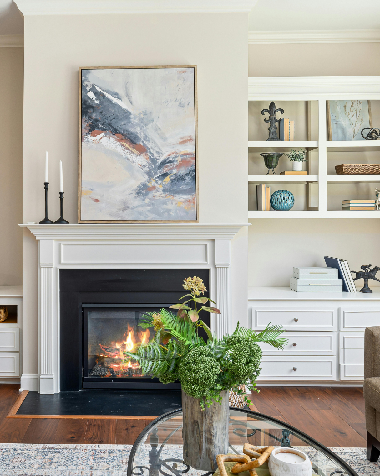 Living room with white fireplace mantel, abstract painting above fireplace, black candlestick holders, built-in white bookshelves with decorative items, and a potted plant on a glass table in front of the fireplace.