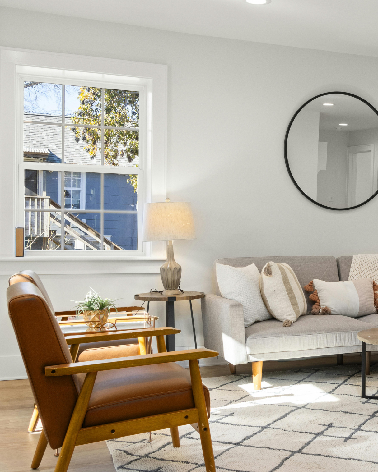 A cozy living room with a large window showing trees and a neighboring house, a beige sofa with multiple pillows, a wooden armchair with a leather cushion, a side table with a lamp, a decorative plant, and a round mirror on the wall.