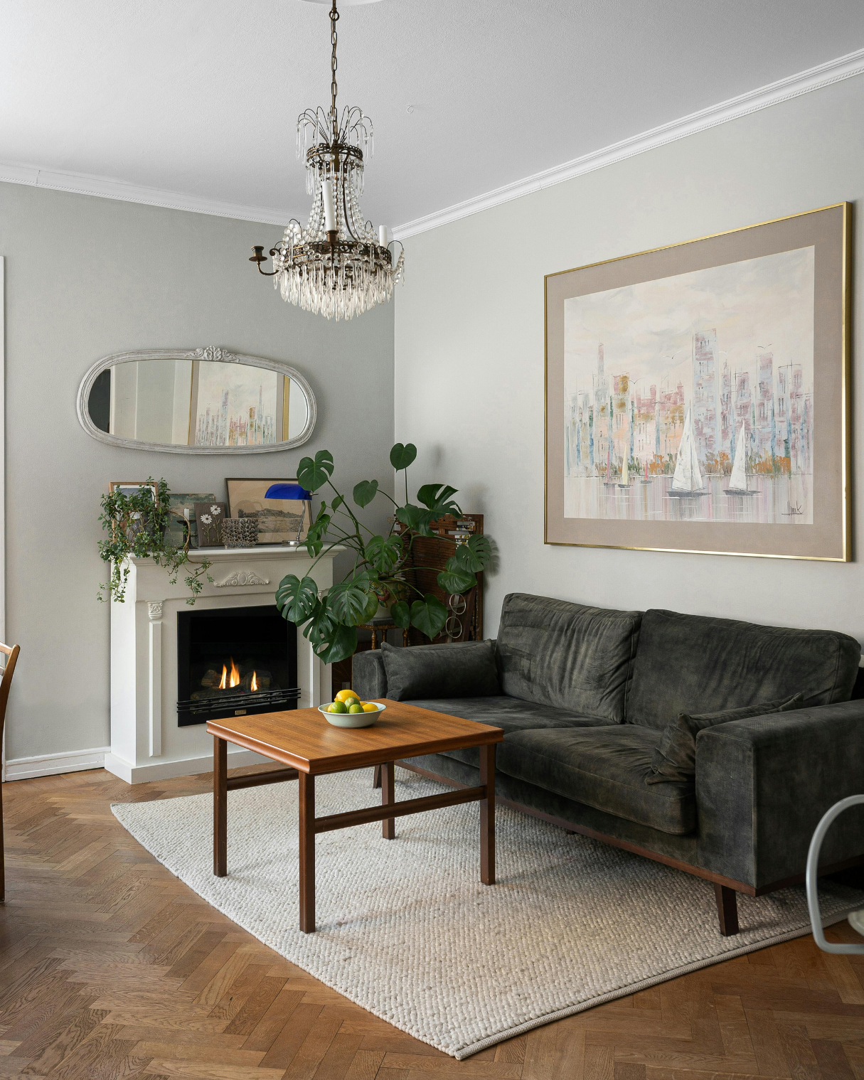 Living room with a gray velvet sofa, a wooden coffee table with a bowl of lemons and limes, a white rug, a fireplace with a mirror above it, a chandelier, a large framed cityscape painting, and green plants.