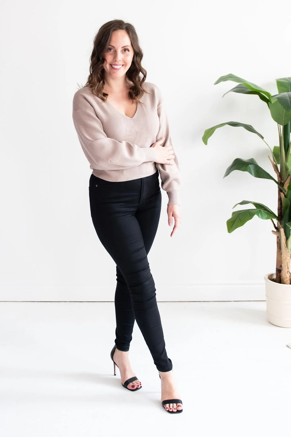 A woman with wavy brown hair smiling and standing with one arm crossed over her waist, wearing a beige sweater, black jeans, and black high heels, next to a potted plant in a white room.