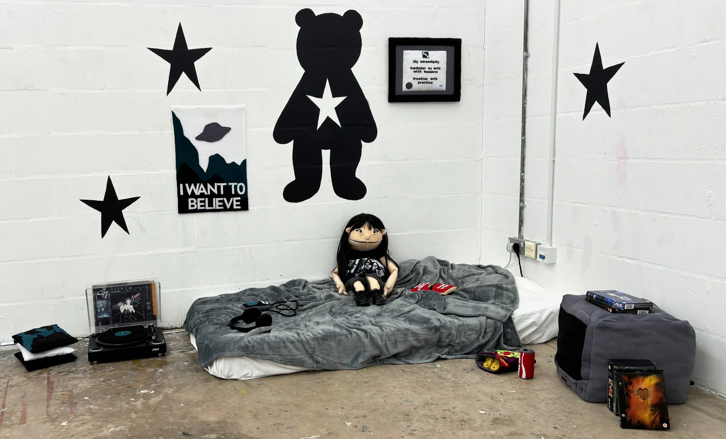 A room with a bed covered in a gray blanket, a plush doll sitting on it, surrounded by various items including a turntable with headphones. The white wall behind has black star and bear-shaped decorations, a framed certificate, and a poster that reads 'I WANT TO BELIEVE' with an image of a mountain and a UFO.