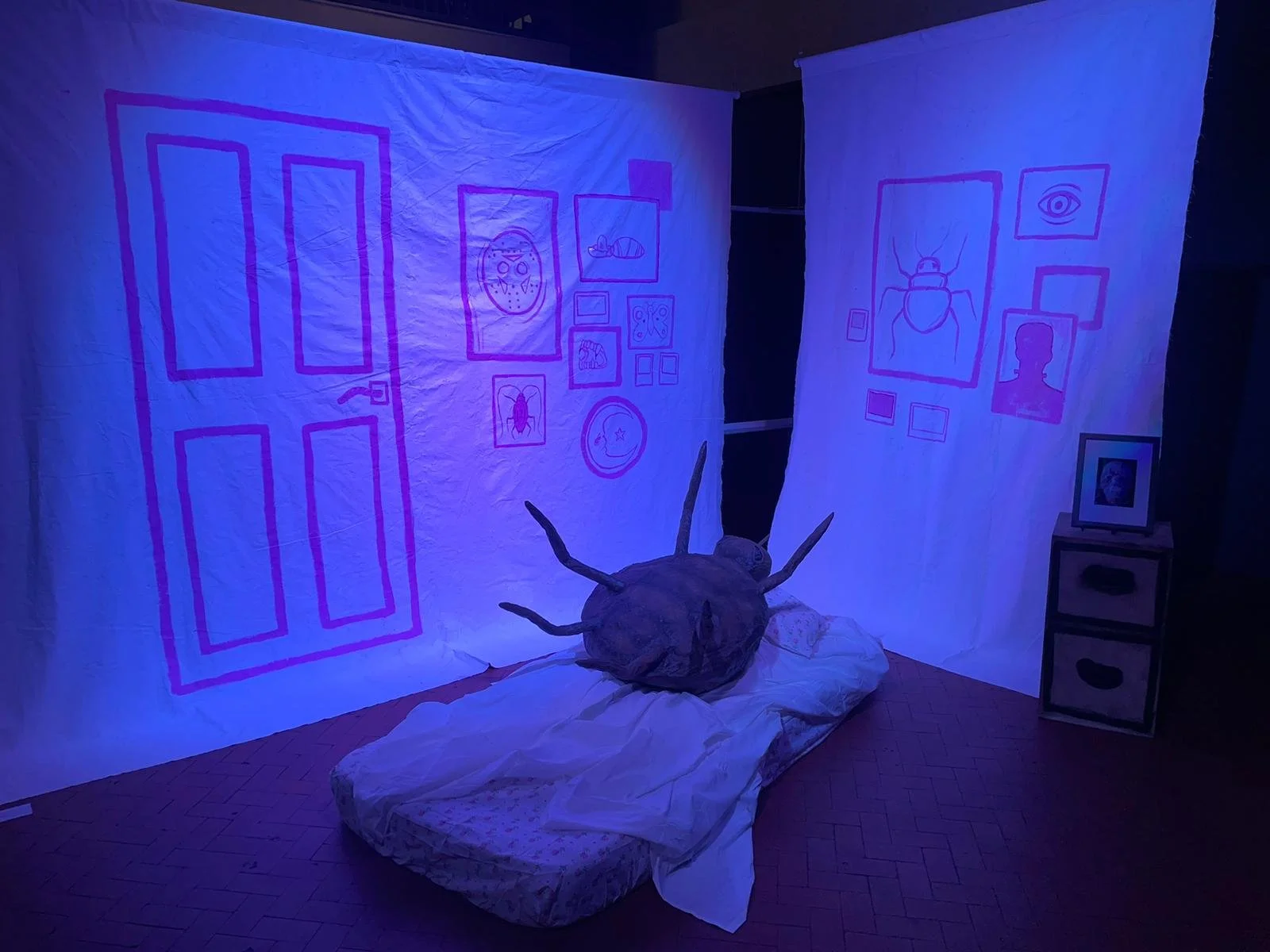 An art installation with pink outline drawings on white fabric walls, featuring various images like a door, a face with a mask, a bug, and walls with frames. In the center, a large bug-like creature sculpture rests on a mattress.