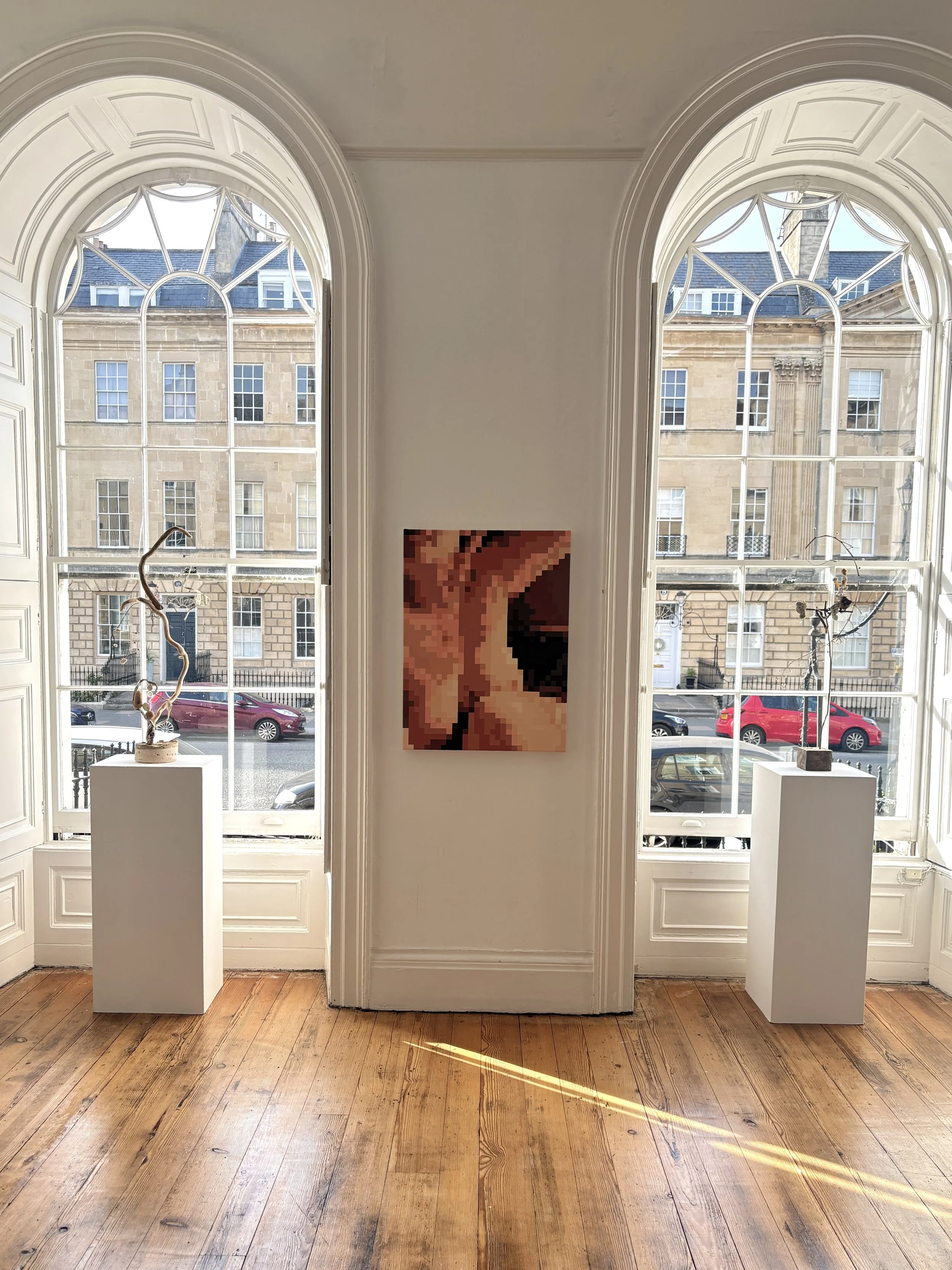 A room with two large arched windows looking onto a street with parked cars and historic buildings. Inside, there are two white pedestals with sculptures, and a pixelated painting hangs on the wall between the windows. The wooden floor has visible grain and natural tones.