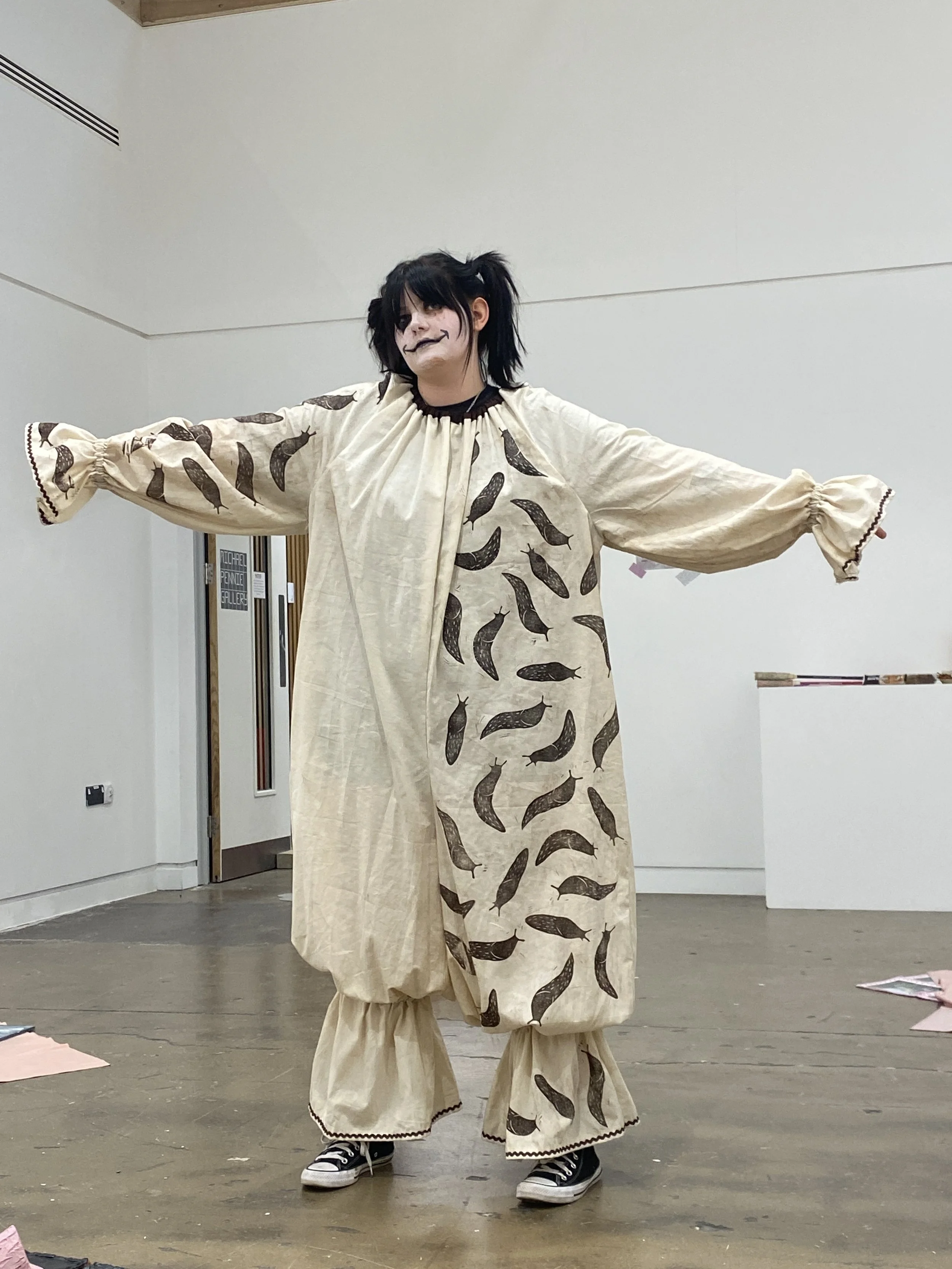Person wearing a clown costume with brown slug print, black and white sneakers, black and white face makeup, standing with arms outstretched in an indoor space.