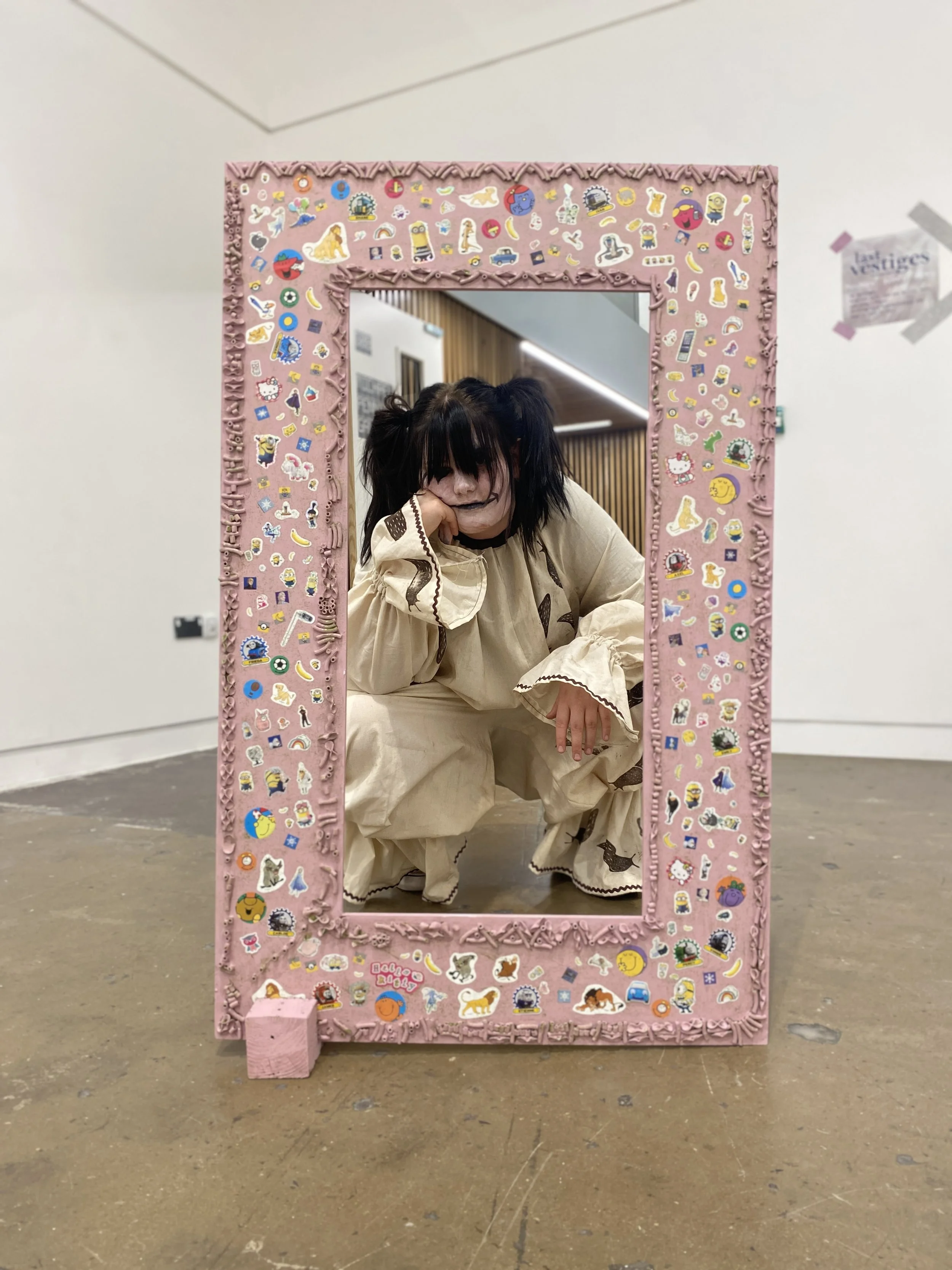 Person with dark hair, dressed in a beige outfit, sitting behind a pink decorated frame inside an art gallery. The frame is adorned with colorful stickers. The person is resting their chin on their hand and looking at the camera.