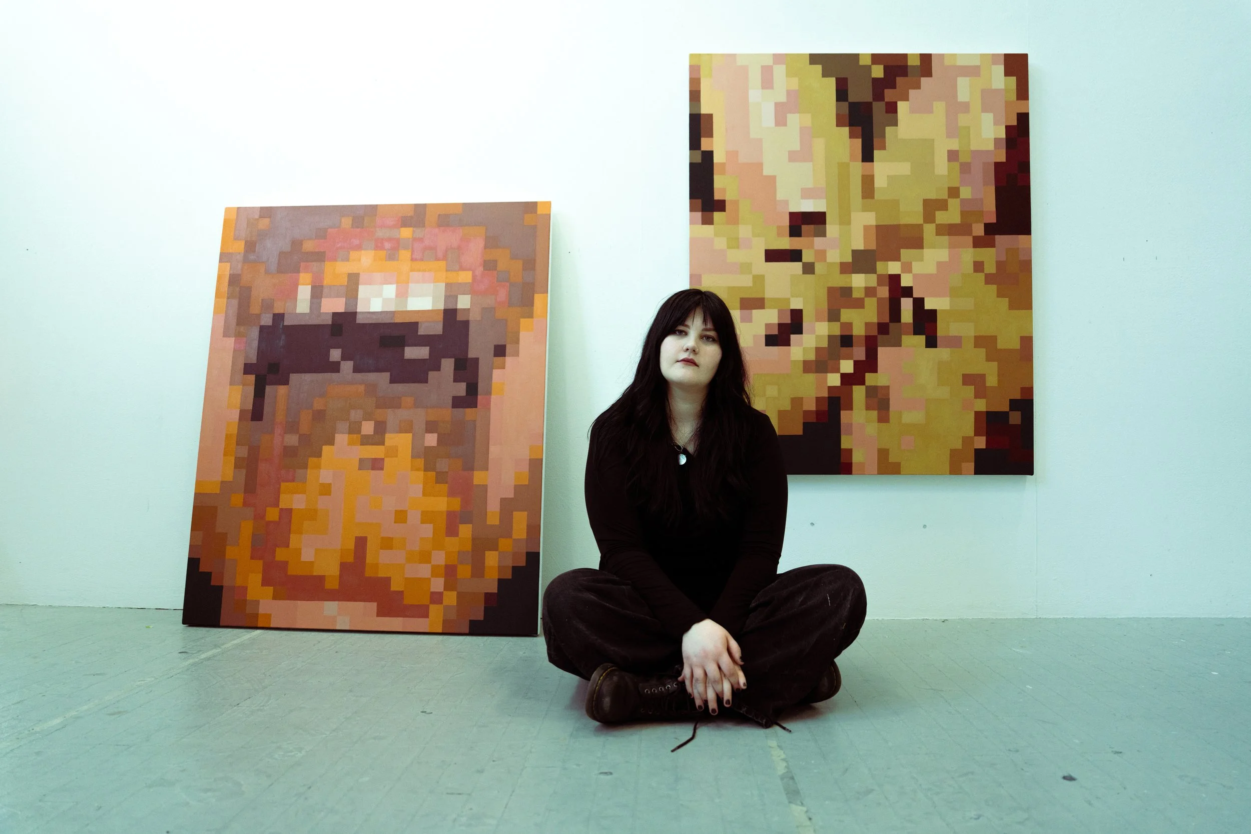 A woman with dark hair and wearing dark clothing sits on the floor with her legs crossed in front of two pixelated art pieces on a light-colored wall.