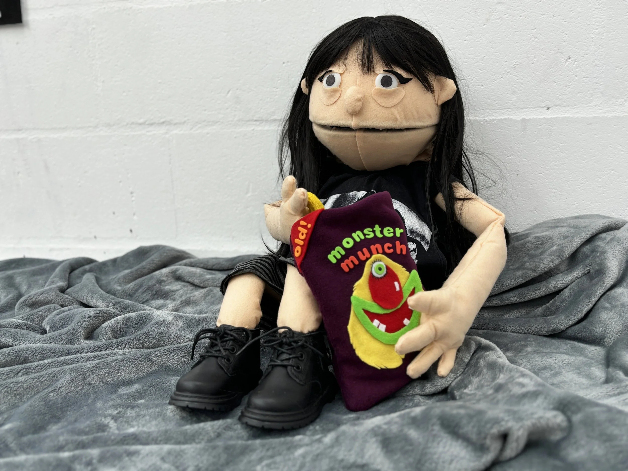 A puppet with black hair sits against a white wall. It is holding a pack of "monster munch" crisps. The puppet is wearing black shoes and a black shirt, and is sitting on a gray blanket.