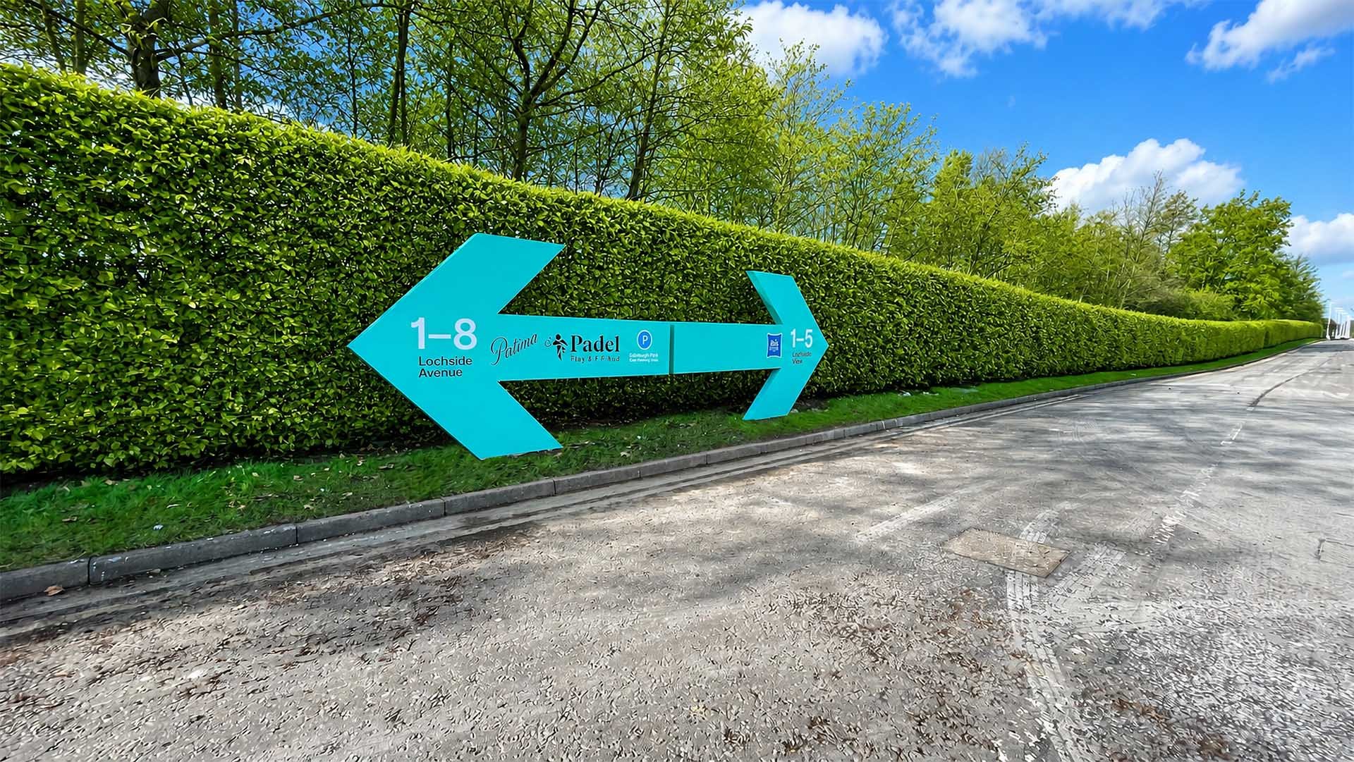 Large, bright teal powder-coated directional arrow signs pointing left and right stand along a lush green hedge and paved road at Edinburgh Park.
