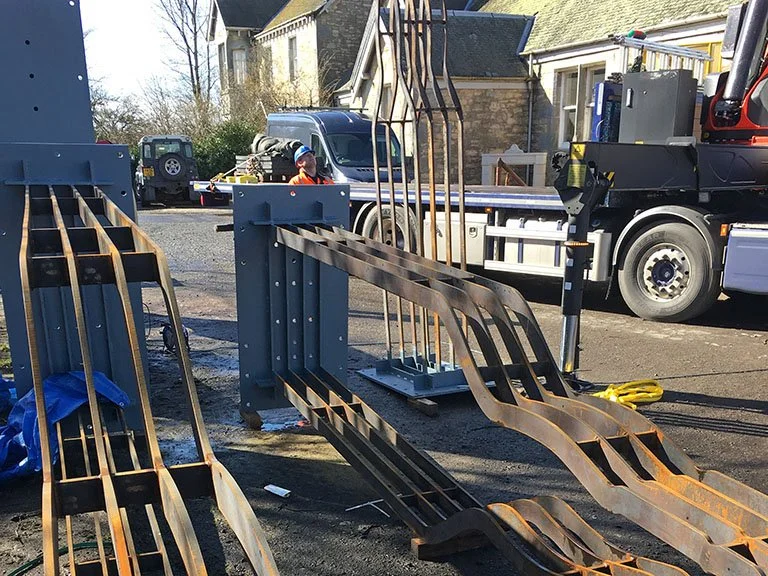Corten steel whisky bottle sculptures lying on the ground next to a flatbed truck fitted with a crane arm, with a worker in a hard hat overseeing loading.