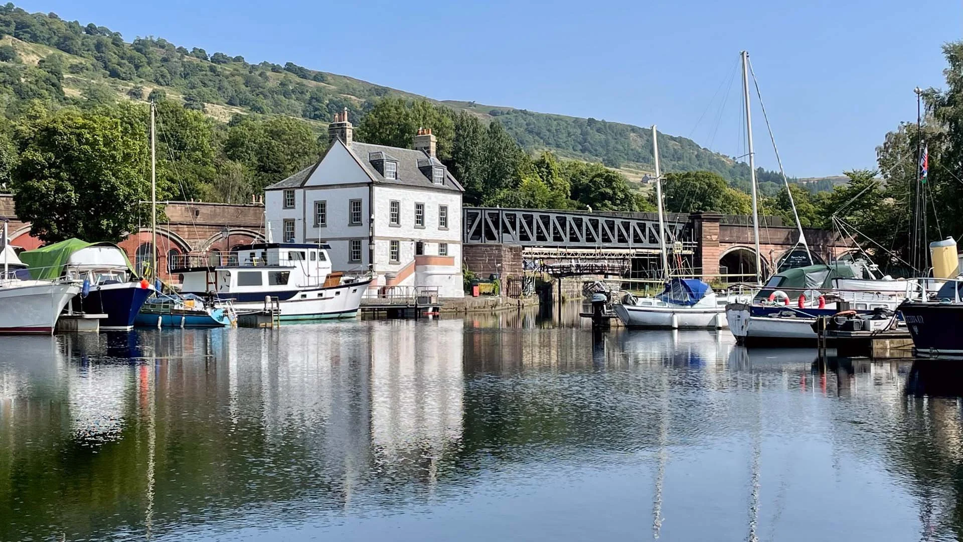 The historic white Customs House sits along the waterfront at Bowling Harbour, surrounded by moored boats, a railway viaduct, and green hills reflecting in the calm water.