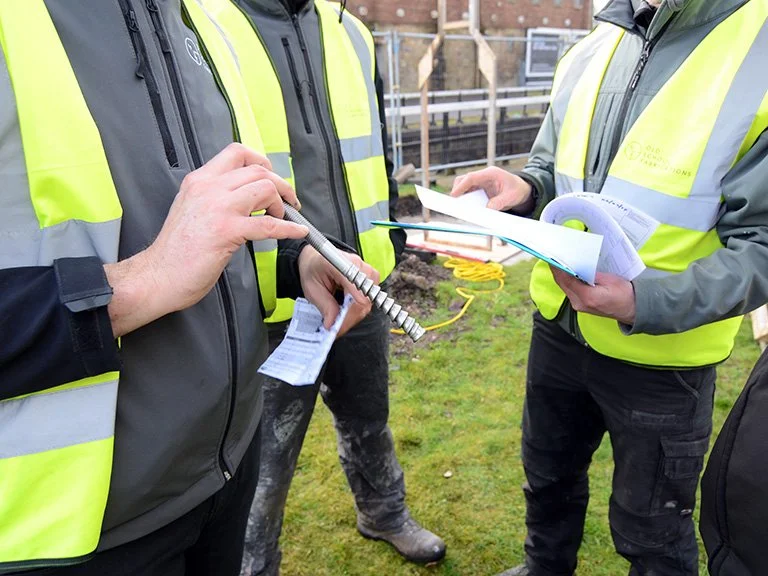 Close-up of workers in high-visibility vests holding large metal ground screw fixings and reviewing plans at Rosebank Distillery.