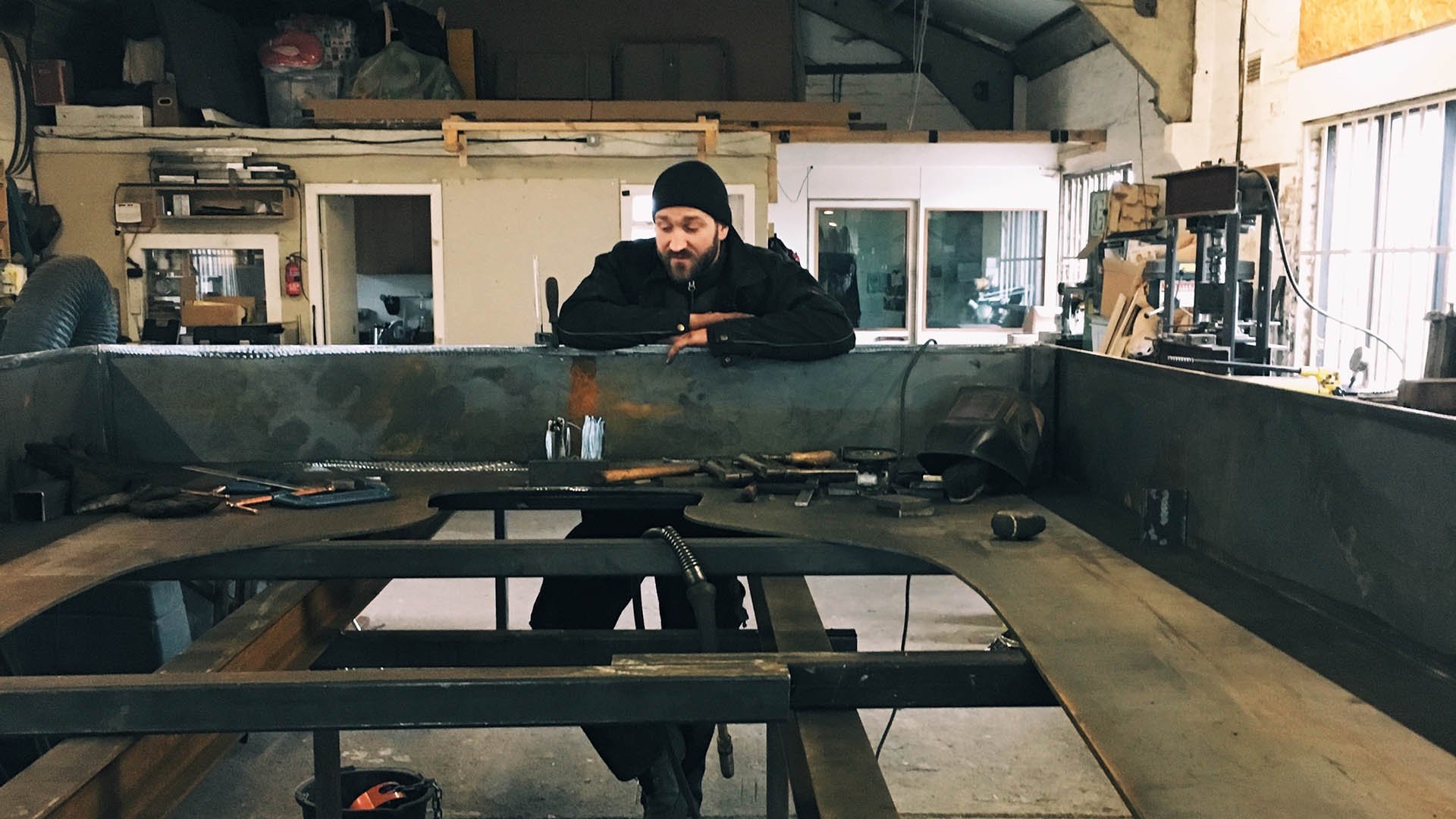 Alt Text Options:  Short & Concise: Fabricator wearing a black beanie leaning over a metal workbench to inspect the progress of the steel vinegar bottle sculpture in a workshop.