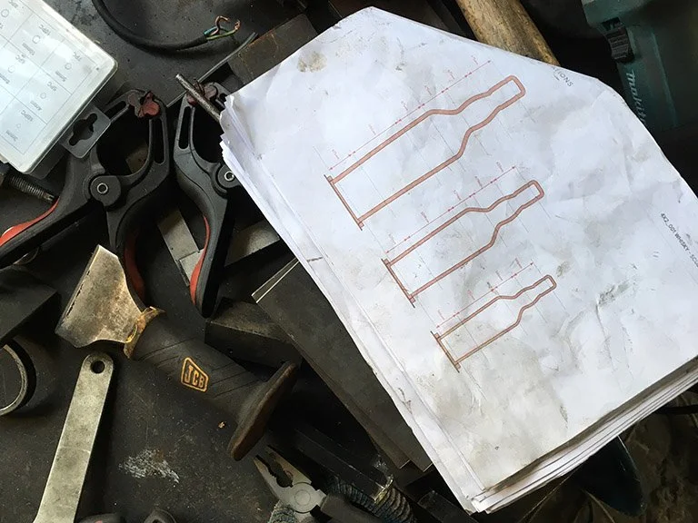 Top-down view of a printed technical blueprint showing whisky bottle sculpture outlines on a workshop bench surrounded by tools.