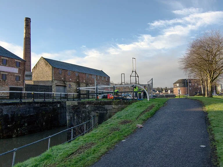 On-site testing of a wooden prototype sculpture for Rosebank Distillery along the canal path, with workers present and historic distillery buildings in the background.