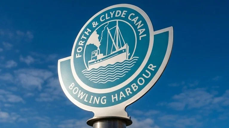 A teal and white circular emblem mounted outdoors against a blue sky features a boat on waves with text reading "FORTH & CLYDE CANAL" around the top and "BOWLING HARBOUR" along the bottom.
