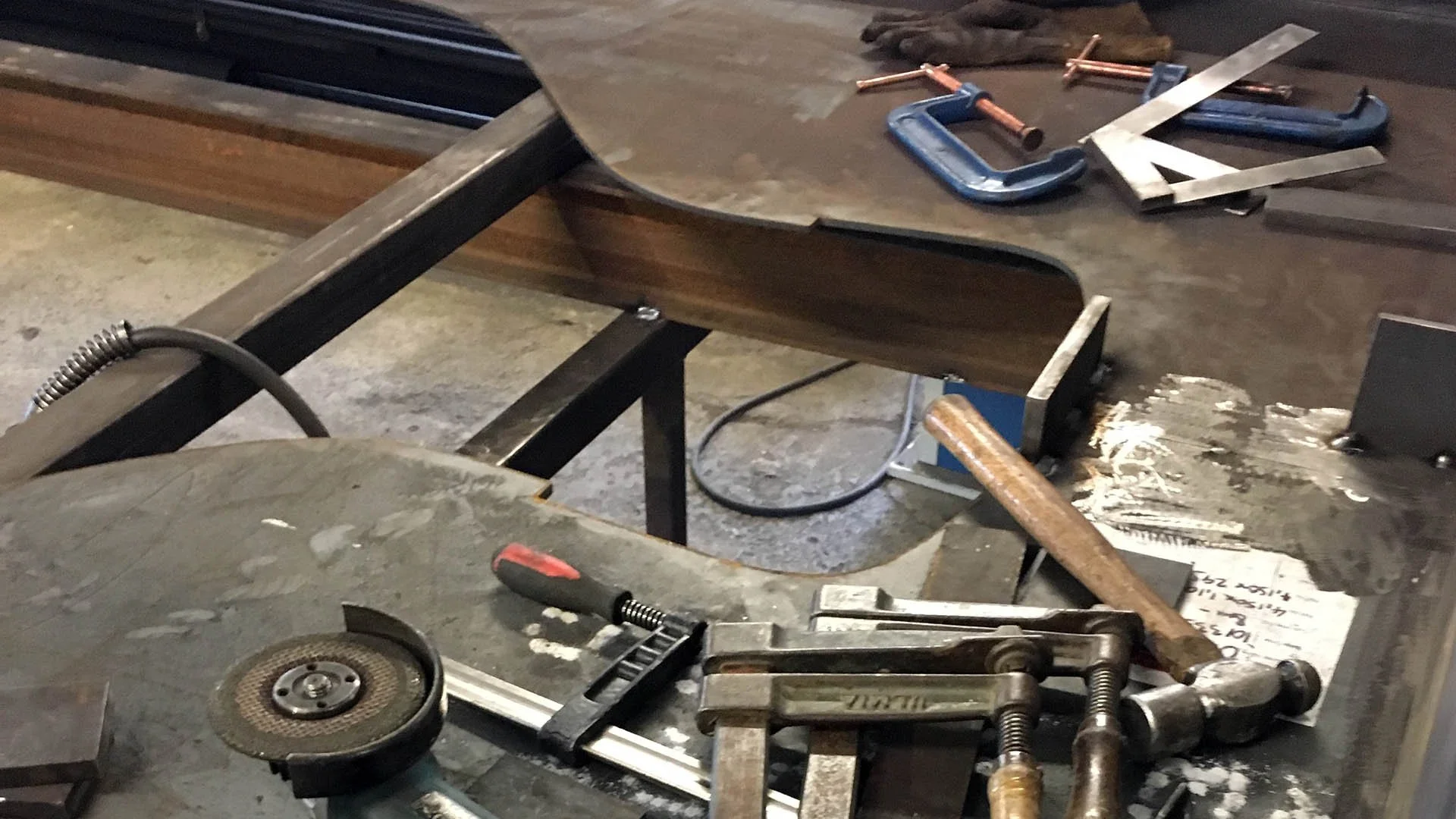 Various metalworking tools including clamps, an angle grinder, and a ball-peen hammer arranged on a steel fabrication table.