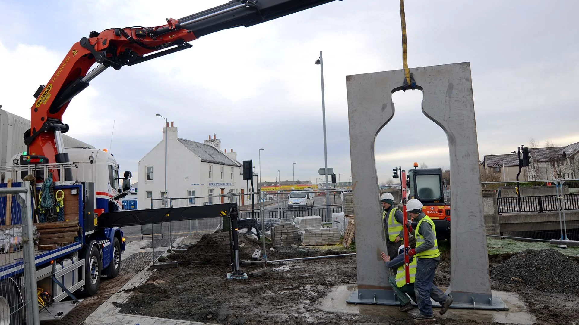 Construction workers using a spirit level as a crane lowers a large metal vinegar bottle sculpture onto its base at Bankside, Falkirk.
