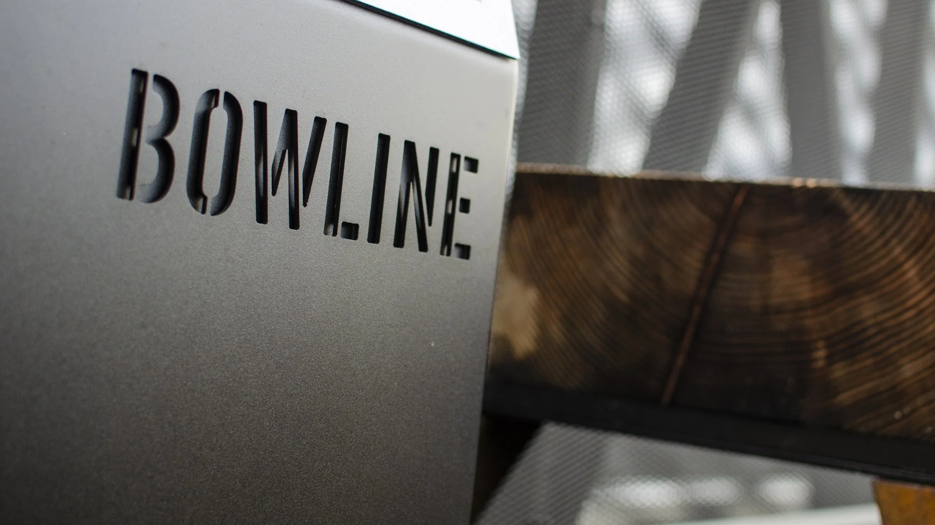 Close-up of metal signage with the word "BOWLINE" cut out in stencil lettering, set alongside a rustic wooden beam.