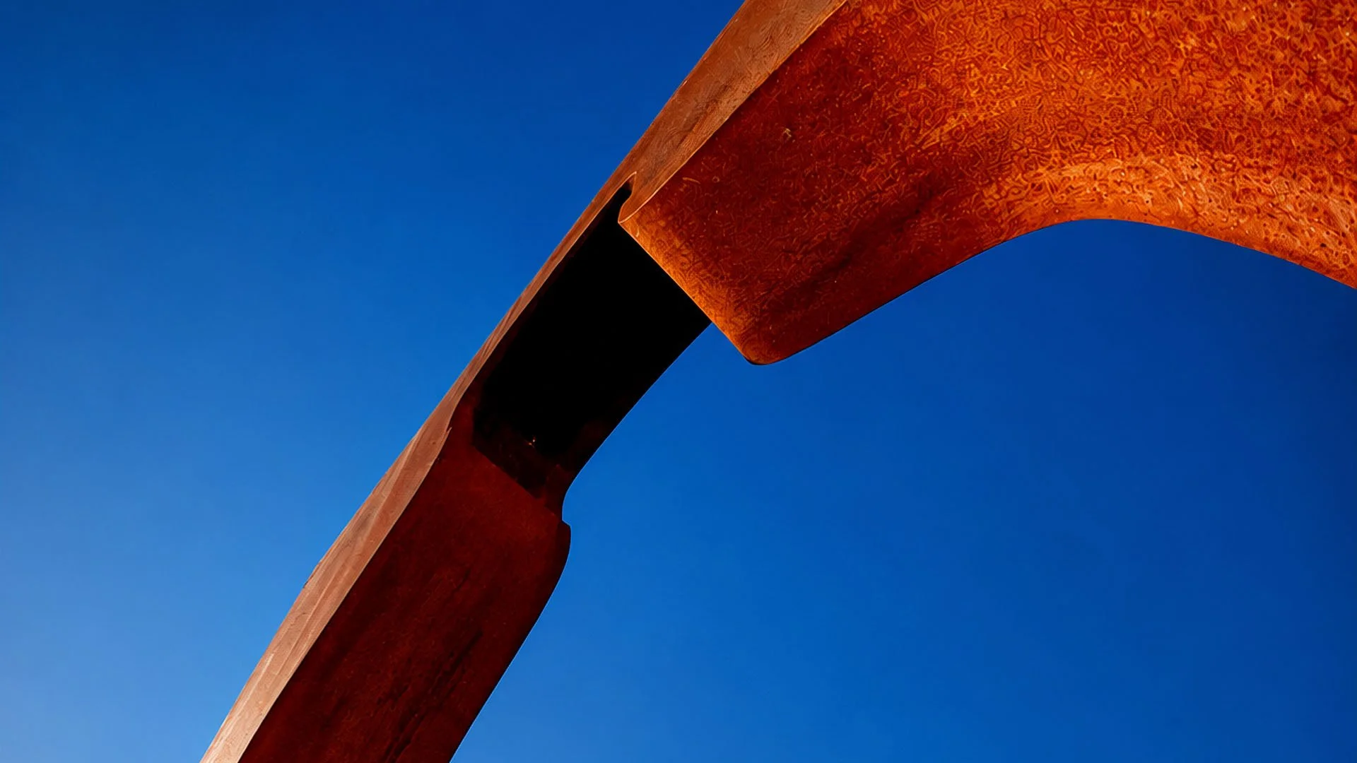 Low-angle view looking directly up from underneath a Corten steel vinegar bottle sculpture against a vivid blue sky.