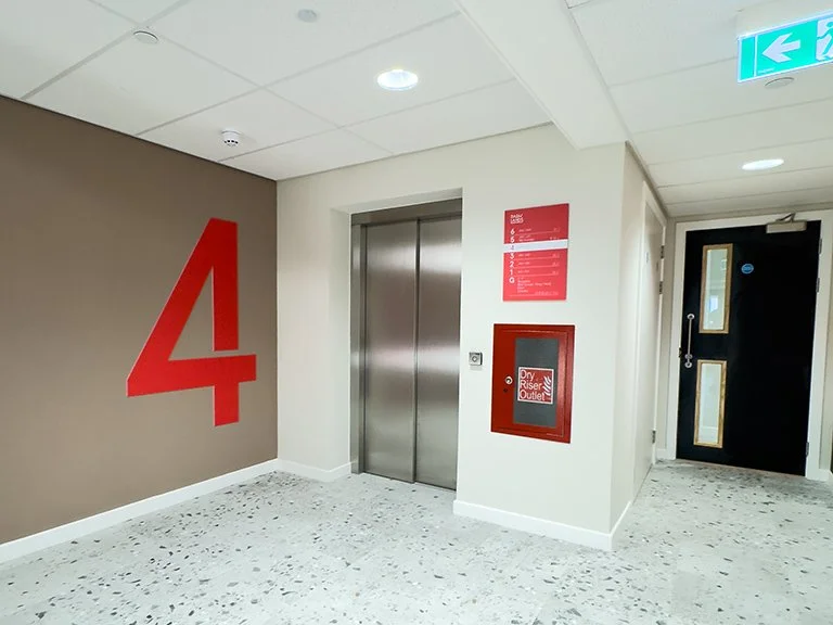 An elevator lobby featuring a large red number "4" painted on a brown accent wall, paired with a red floor directory sign and a metal elevator door above speckled flooring.