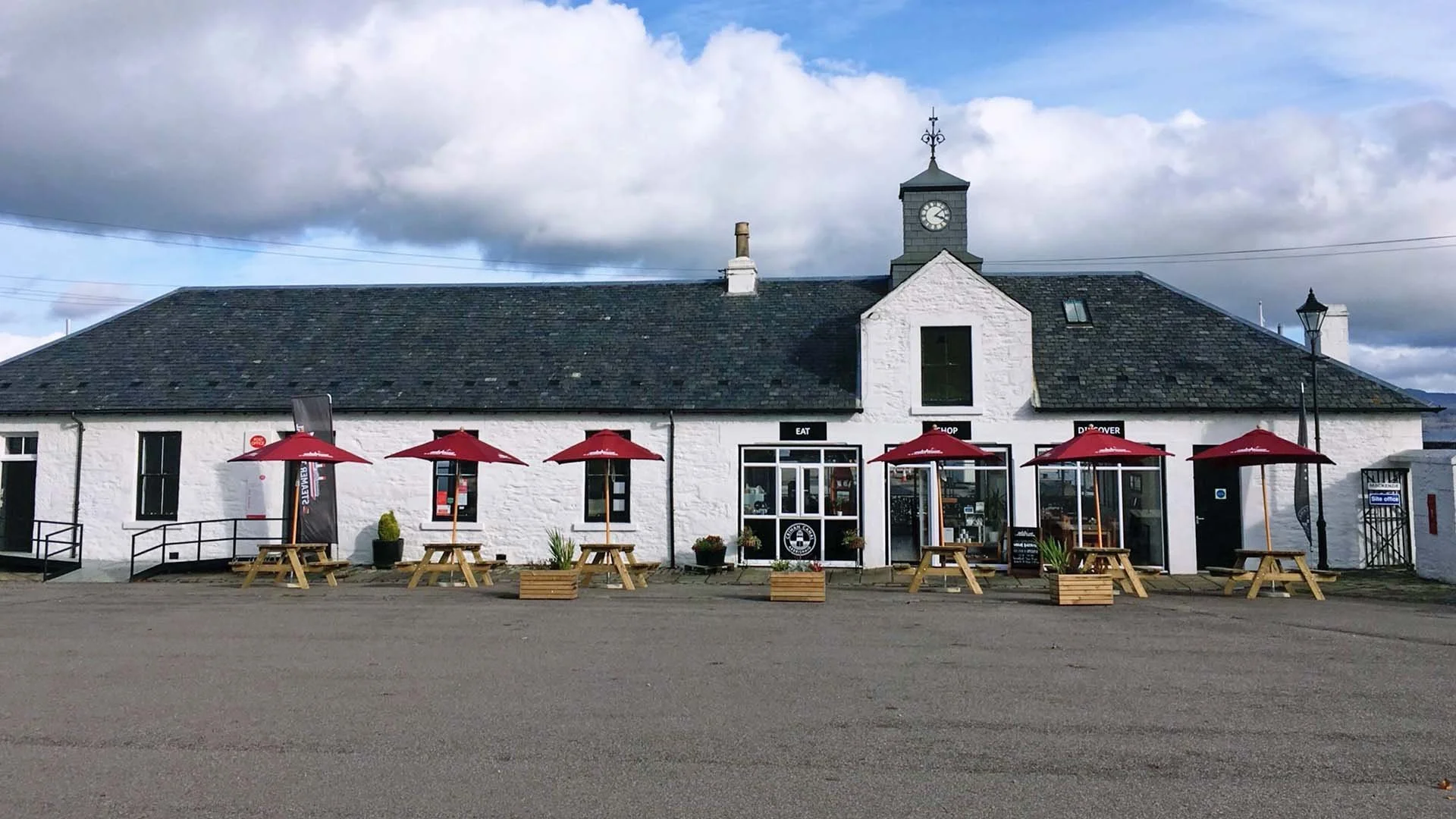 Exterior of The Steamer Terminal at Ardrishaig Harbour, a white single-story stone building with a clock tower and red patio umbrellas over picnic tables.