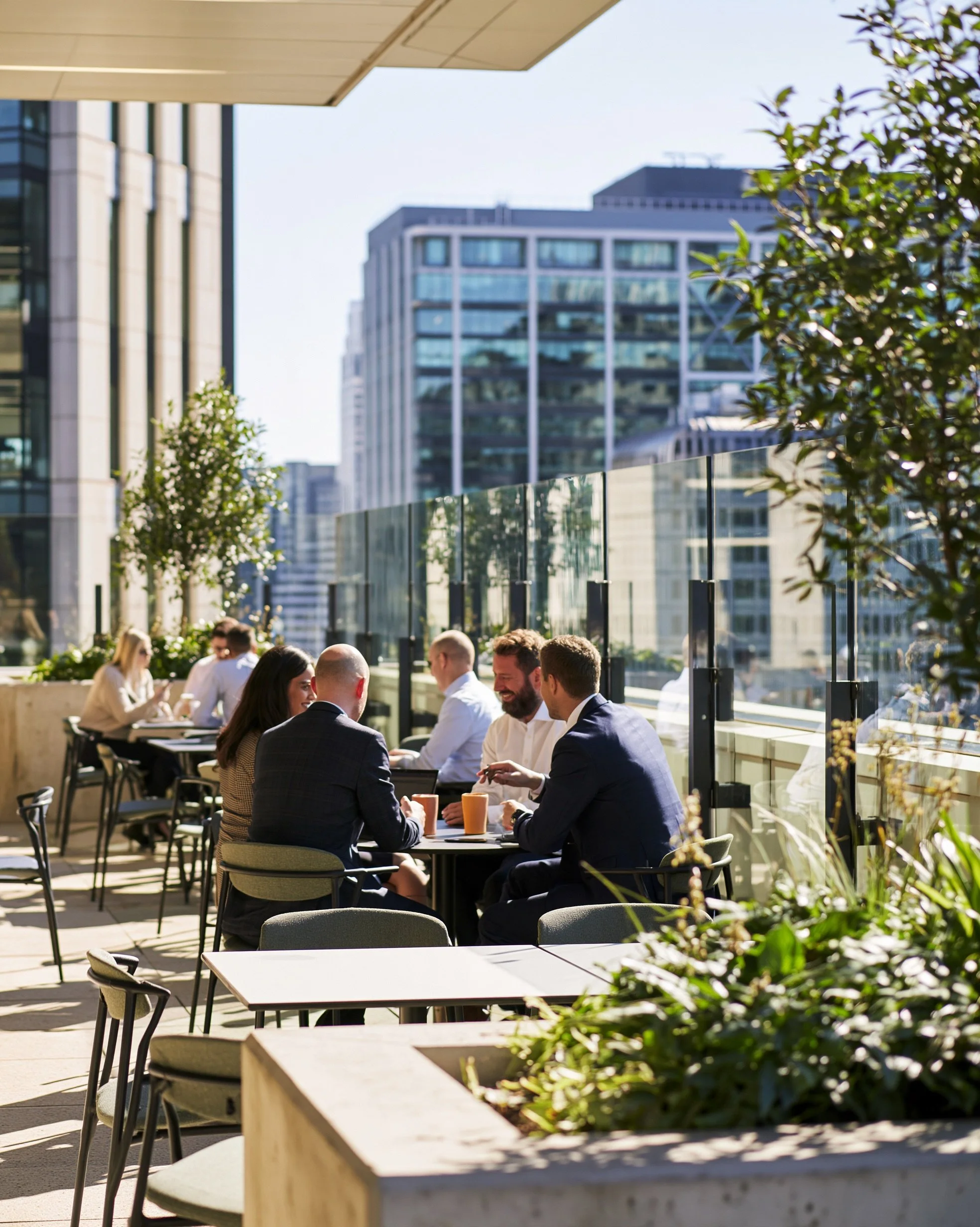 People sitting at outdoor tables on a rooftop terrace, surrounded by glass barriers, with tall office buildings and a clear sky in the background.