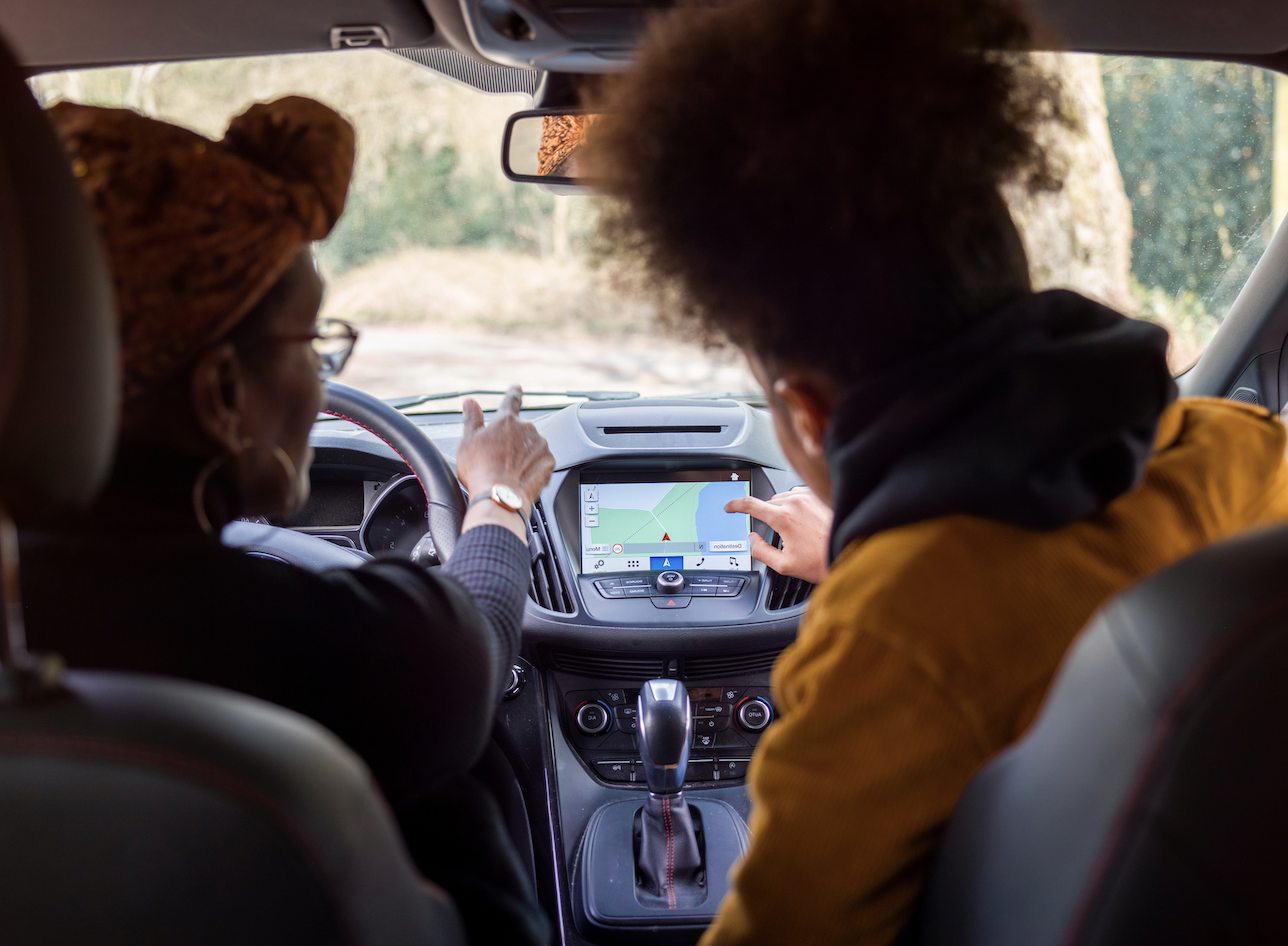 Two people sitting in a car, looking at a GPS navigation screen on the dashboard. One person is driving and the other is pointing at the screen.