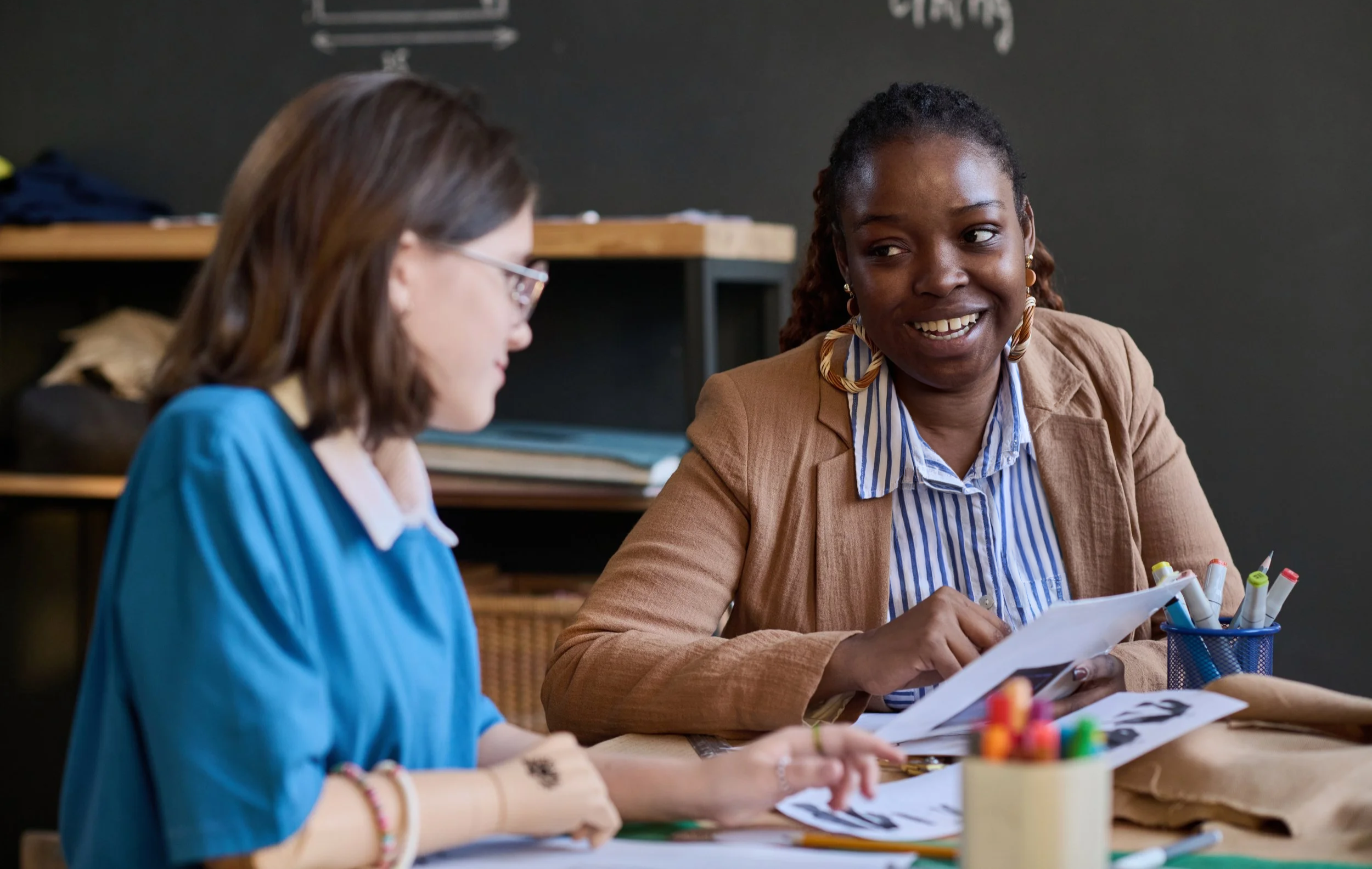 A woman smiling and talking to a girl with glasses in an indoor setting, possibly a classroom or office, with various papers and markers on the table.
