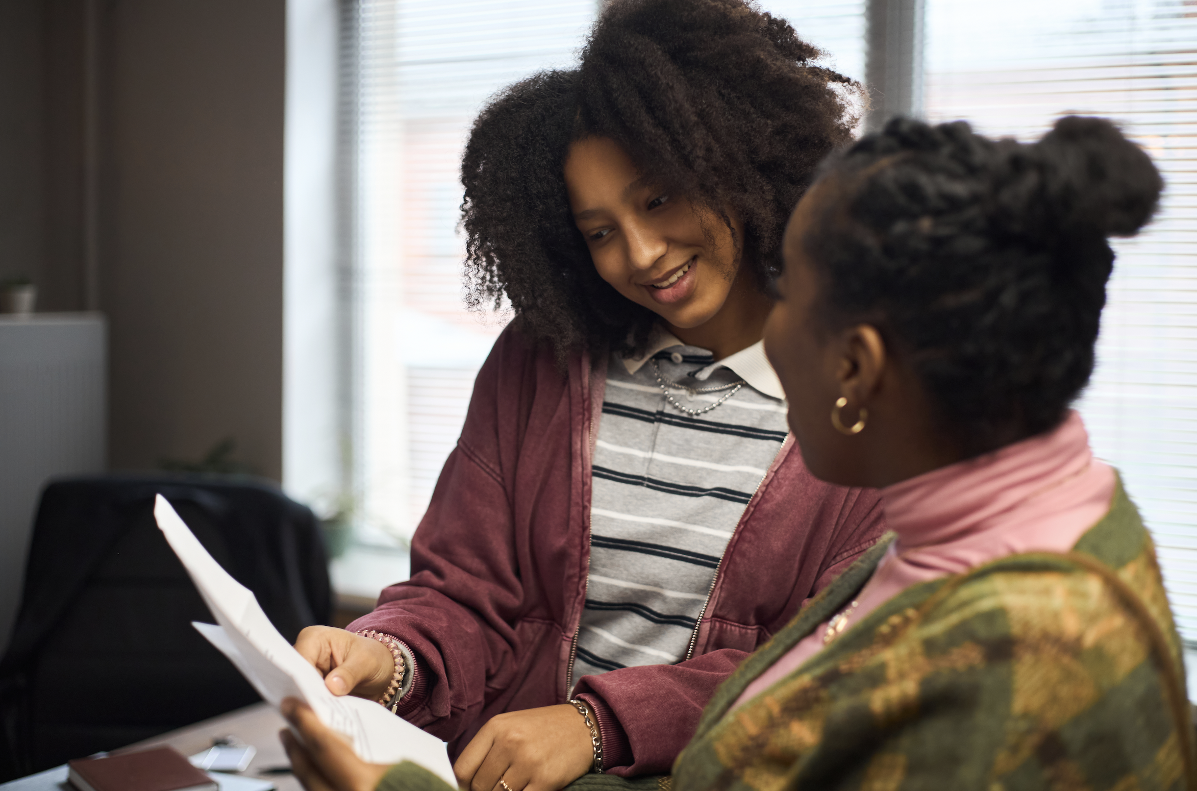 A young woman is talking to a middle-aged woman. They're looking at paperwork in an indoor setting.