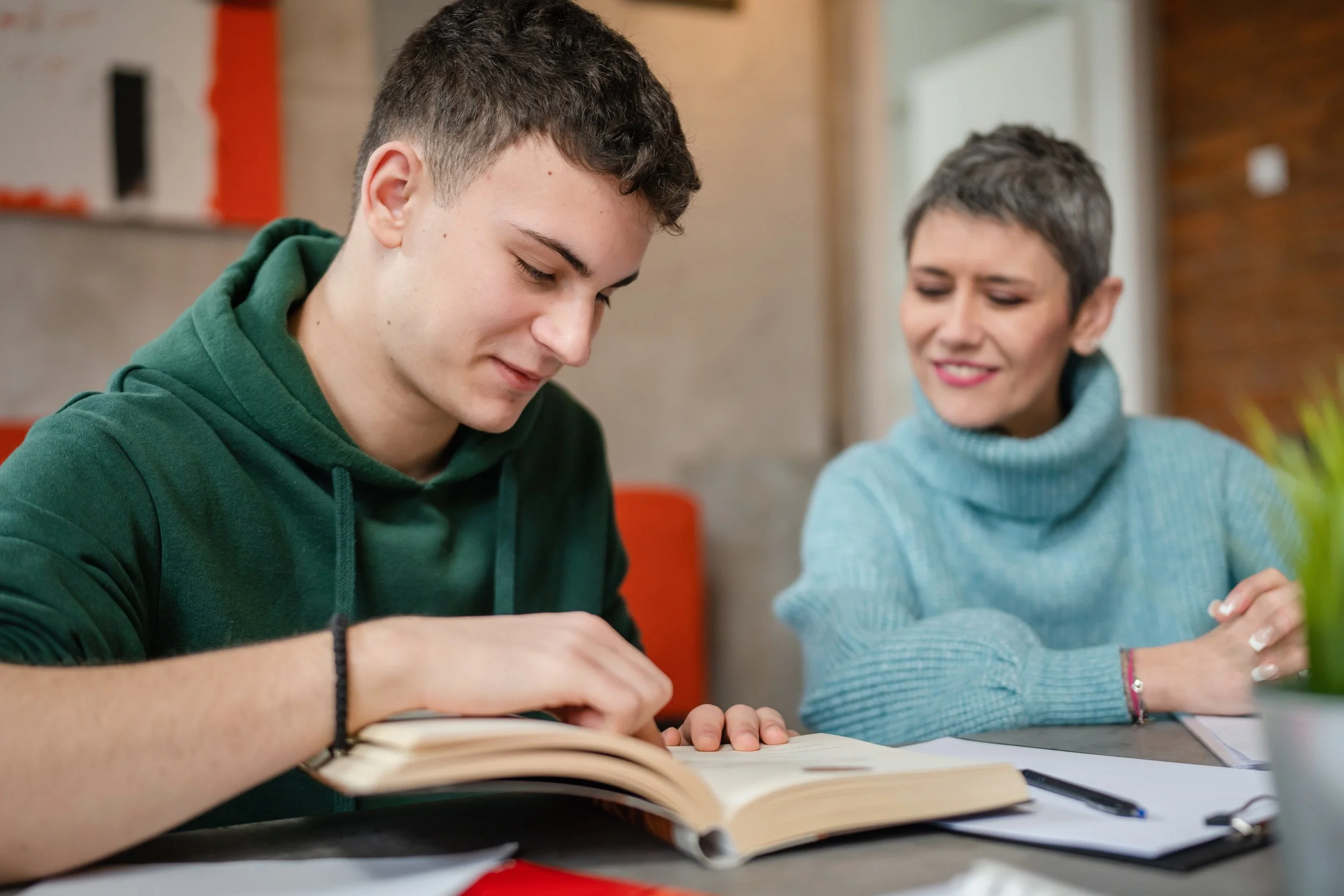 A young man in a green hoodie and a middle-aged woman in a light blue turtleneck sitting at a table reading a book together.