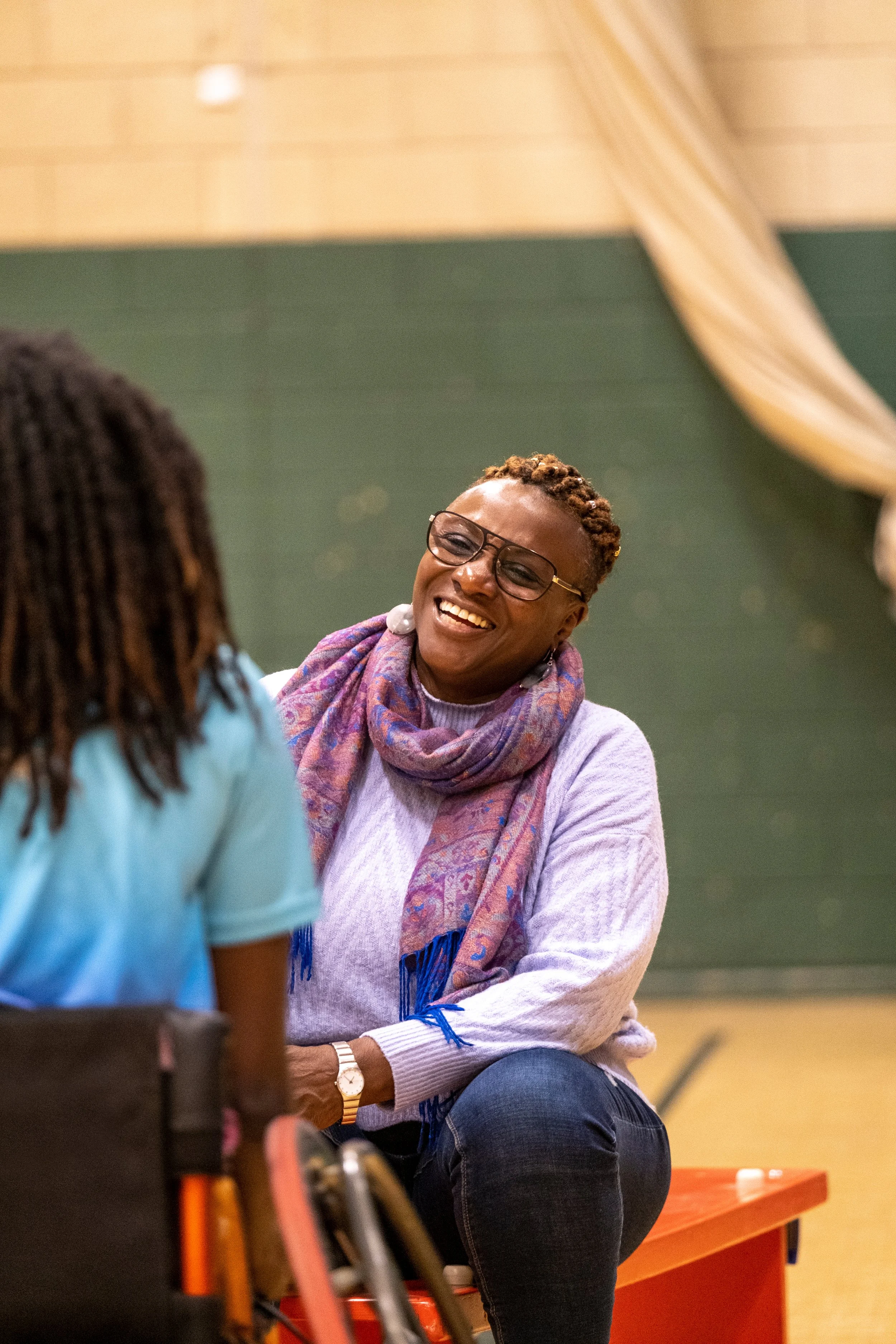 A woman with glasses and short, curly hair smiling and talking to another person in a classroom or community center setting.