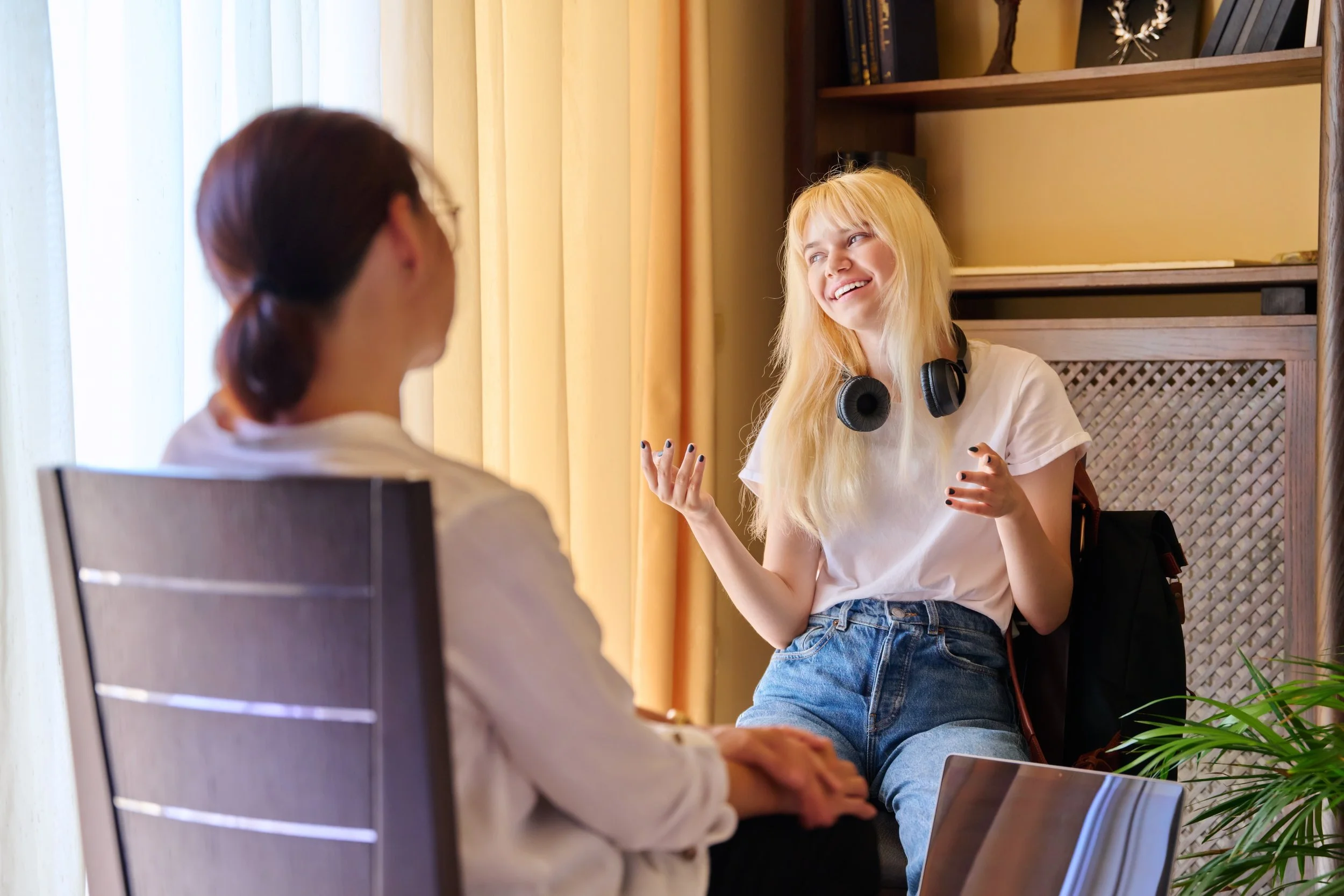 Two women sitting and talking indoors with one woman wearing headphones around her neck and smiling.