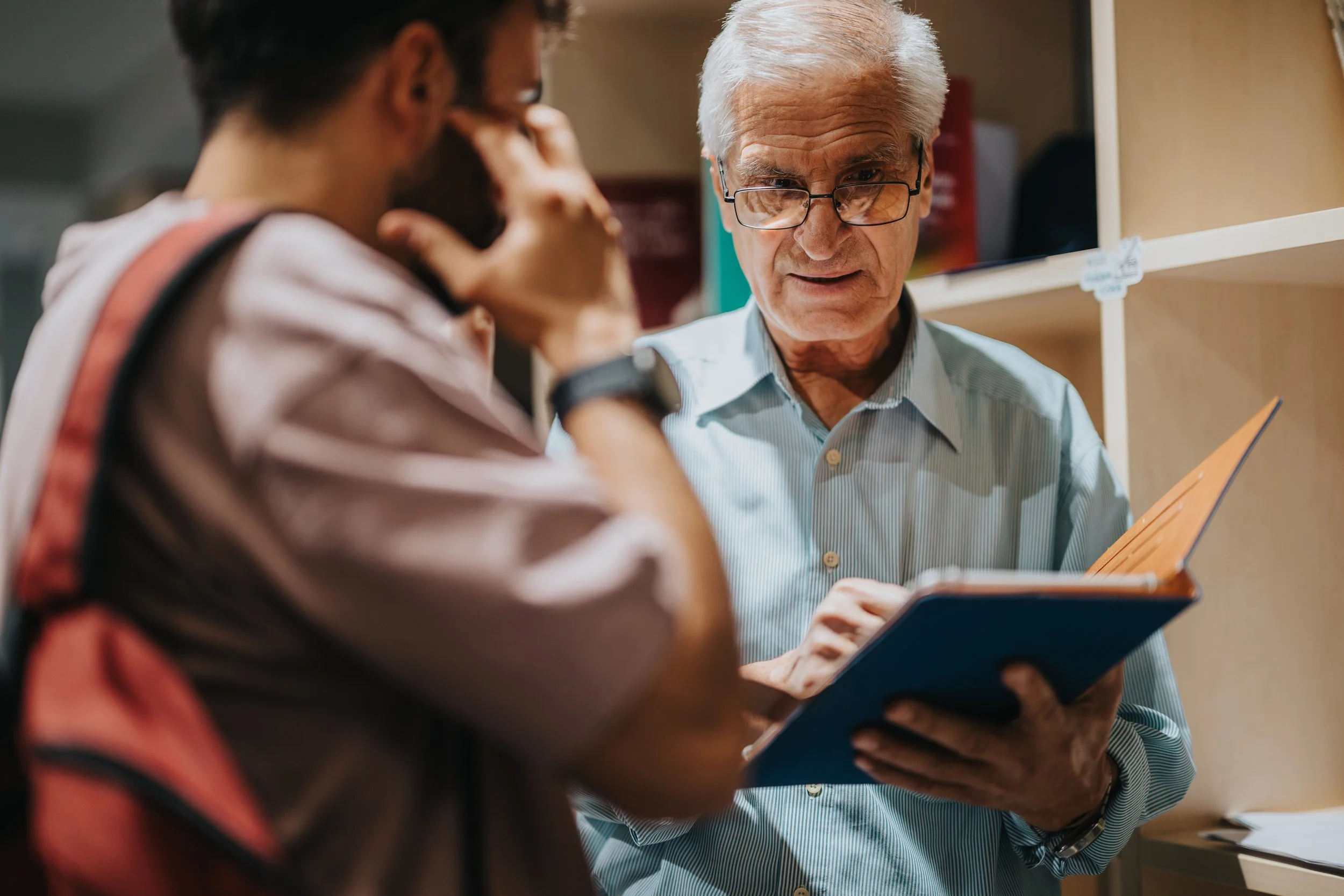 Older man with white hair and glasses showing a young man with dark hair and beard a folder or tablet while they stand near shelves in an indoor setting.