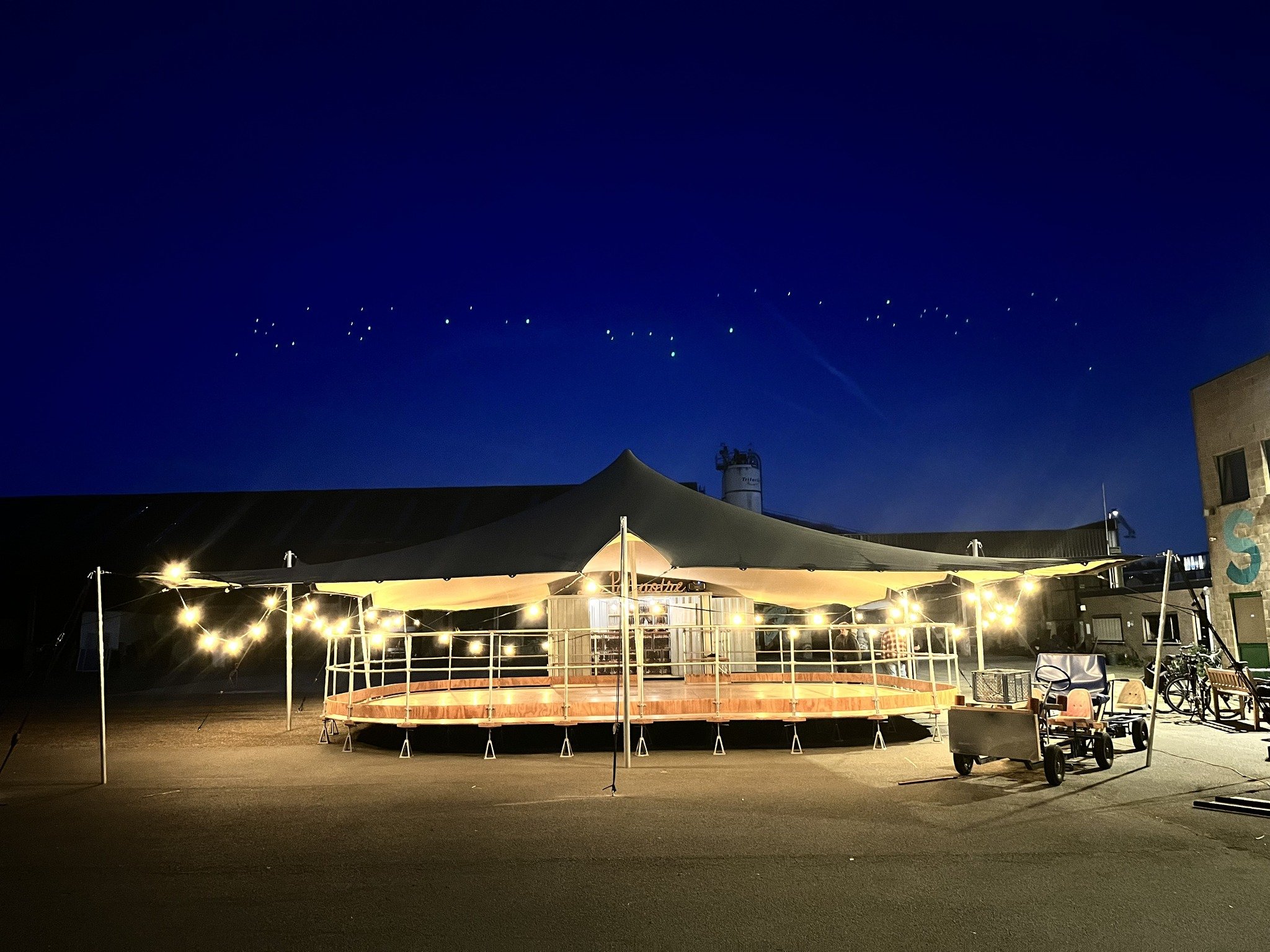 Night scene of outdoor area with string lights, tents, and a stage or platform with fencing. Buildings and equipment are visible in the background under a dark sky.