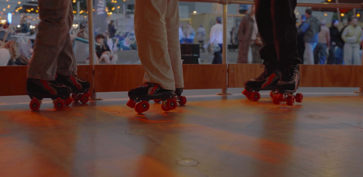 Three people wearing roller skates on an indoor rink, with a crowd of people and festive decorations in the background.