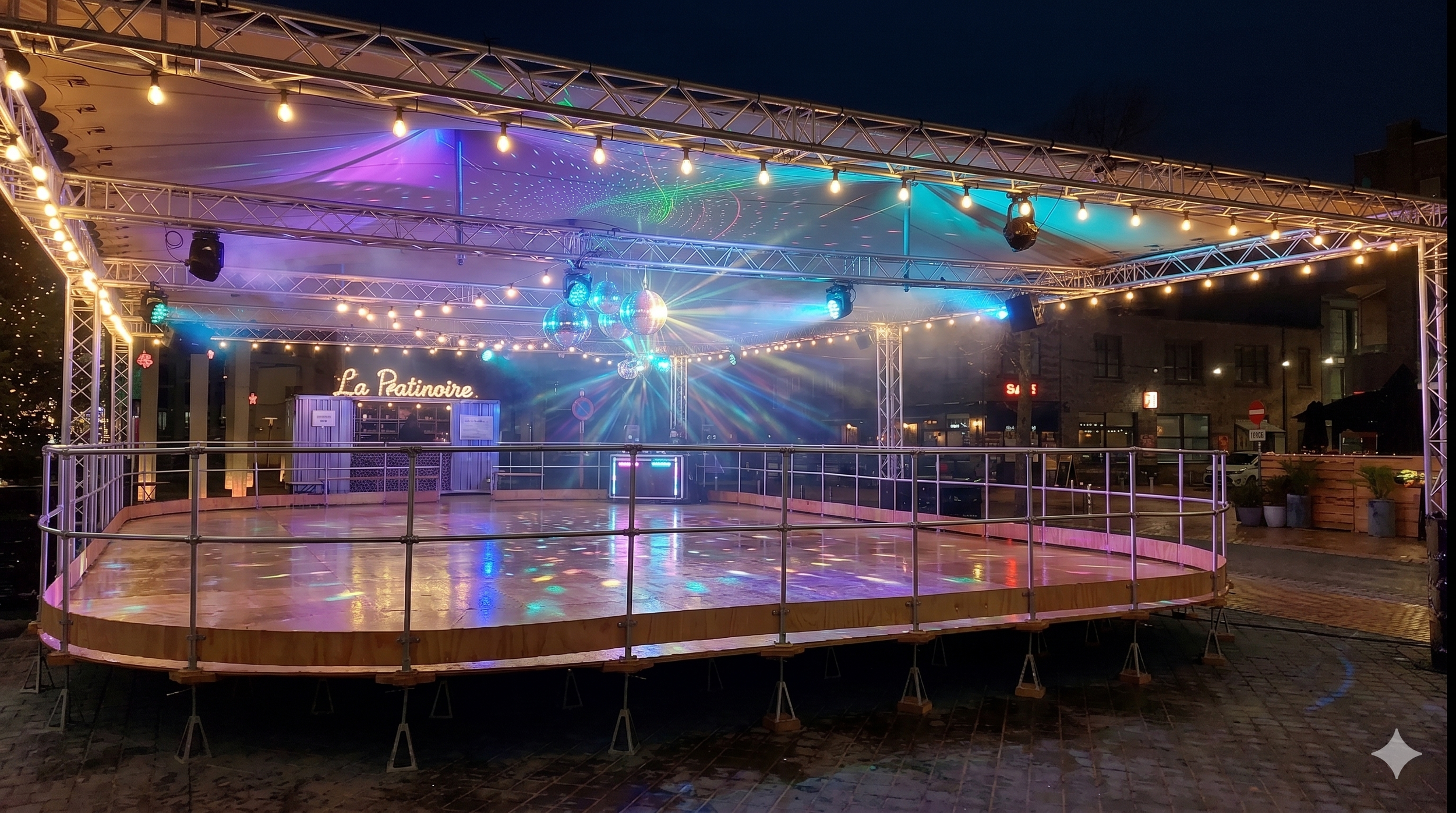 Empty outdoor dance floor with lights and disco balls under a canopy at night, with buildings and signs in the background.