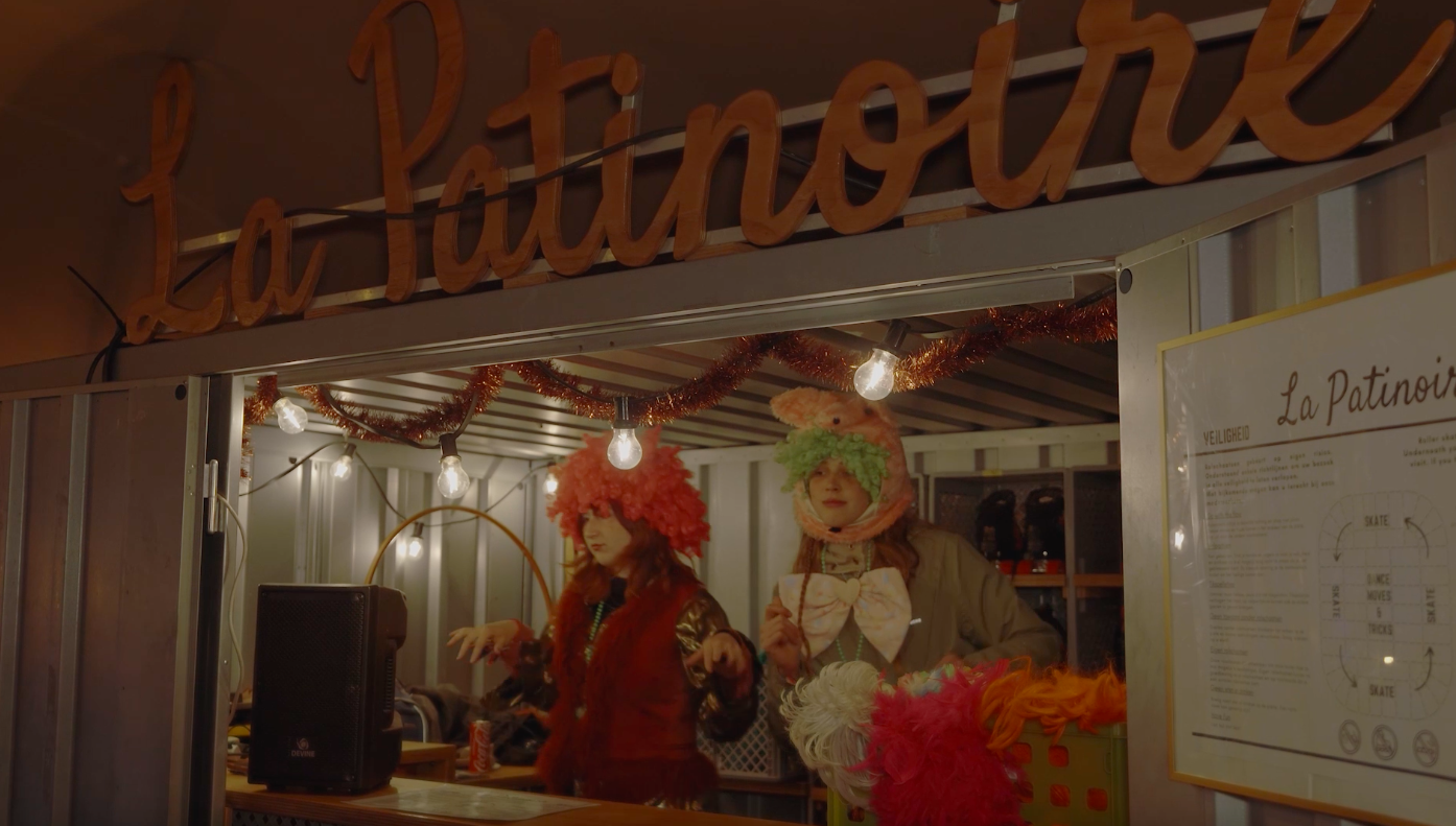 Two women dressed in colorful, fluffy costumes with headpieces resembling animals, standing inside a decorated booth labeled 'La Patinoire,' with string lights and tinsel hanging overhead.